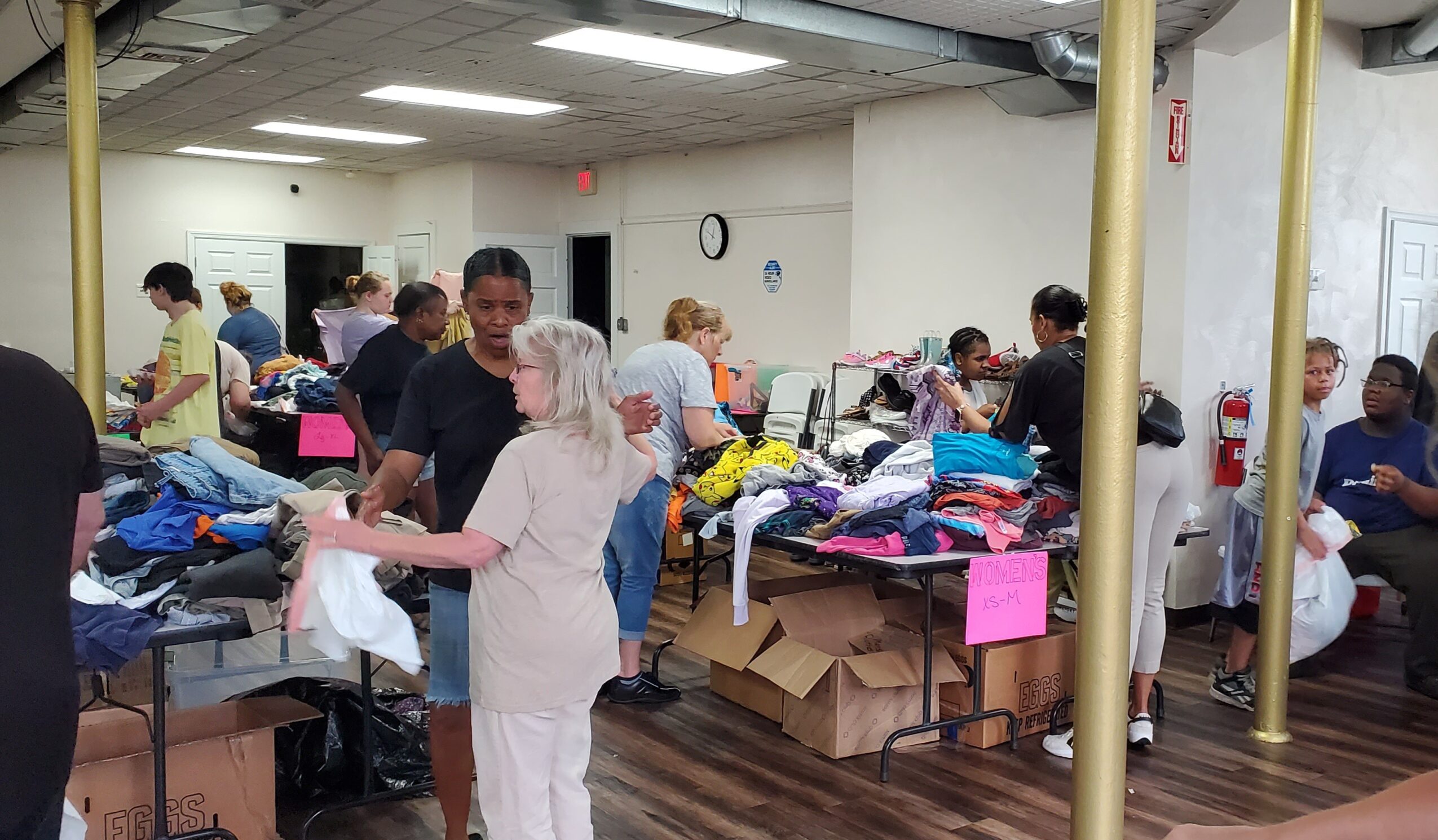 People sorting and organizing clothes on tables in a room with boxes and supplies, some standing and some sitting.