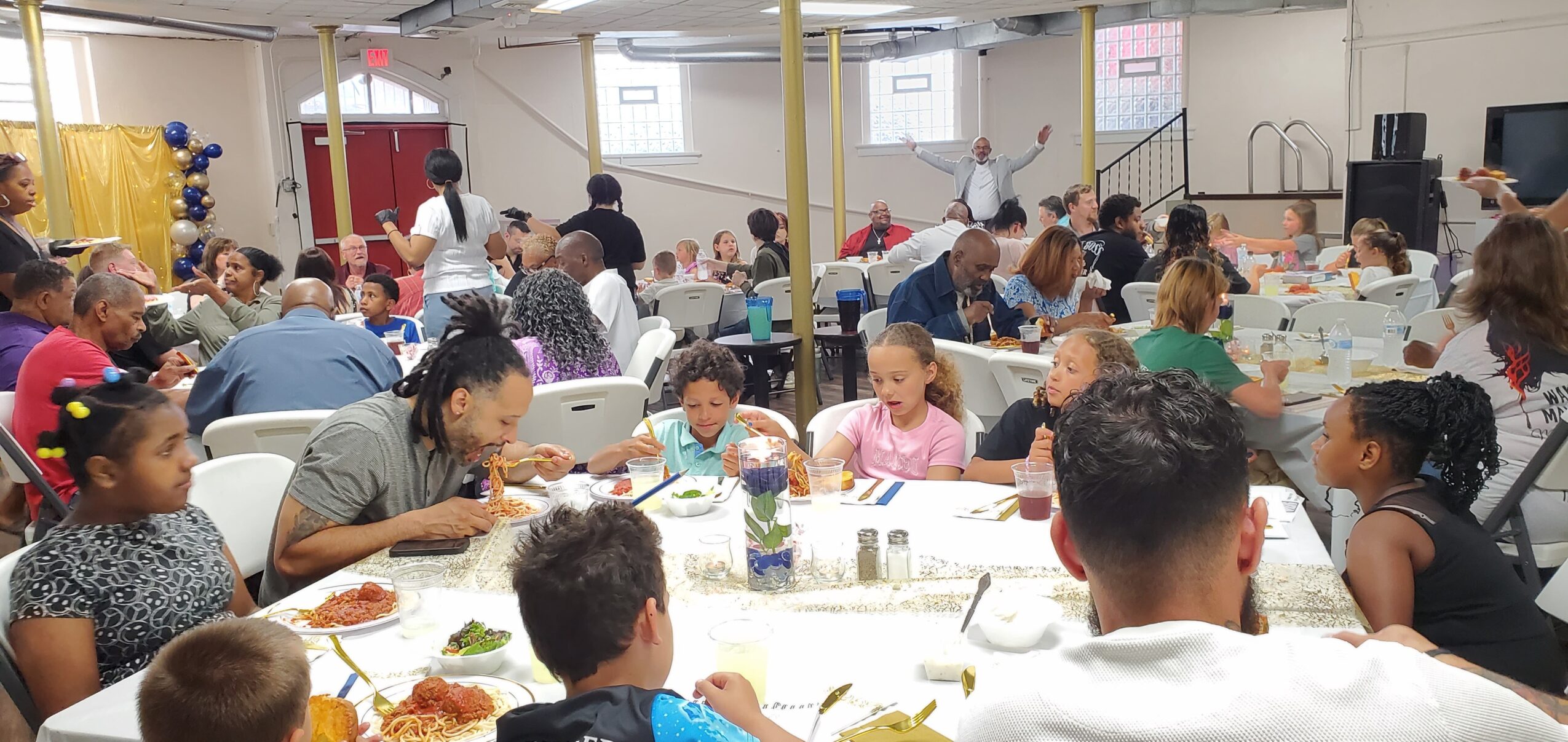 Children and adults sit at tables in a large room, eating and socializing during an event.