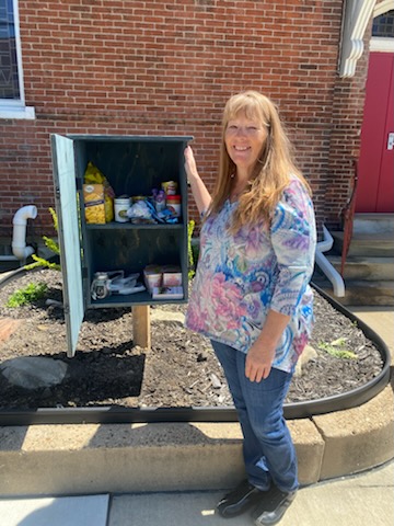 Woman with long hair smiling outdoors next to a small blue cabinet with food items, in front of a brick building.