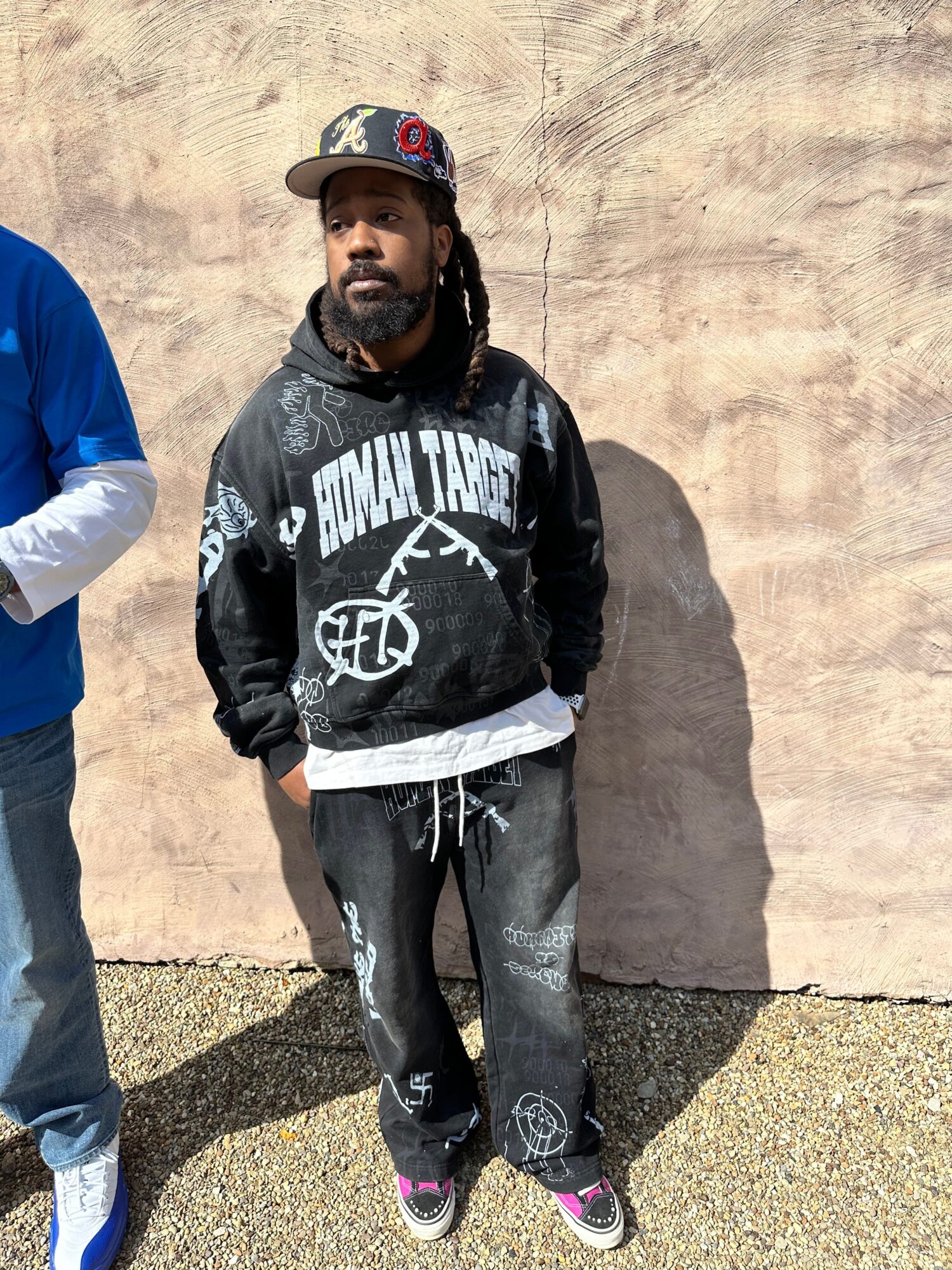 Person standing outdoors against a beige wall, wearing a black jacket, black pants, and a cap, with a shadow on the ground.