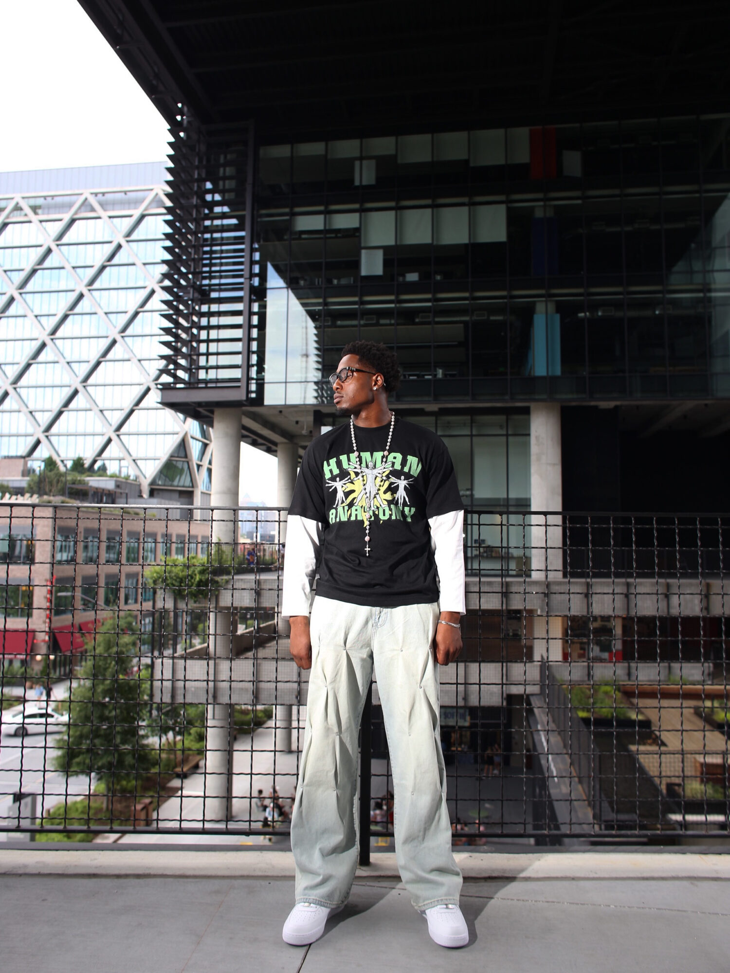 Young man standing outdoors in front of modern building, wearing black hoodie, white pants, and white shoes.