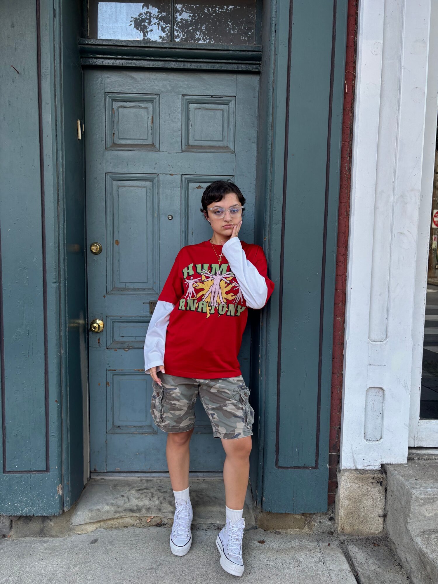 Person standing in front of a closed door, wearing a red and white shirt, camouflage shorts, and white shoes.