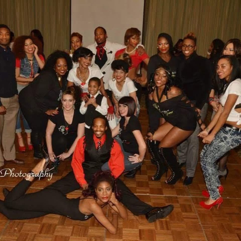 Group of people posing together indoors, some standing and some sitting or lying on the floor, smiling and looking at the camera.