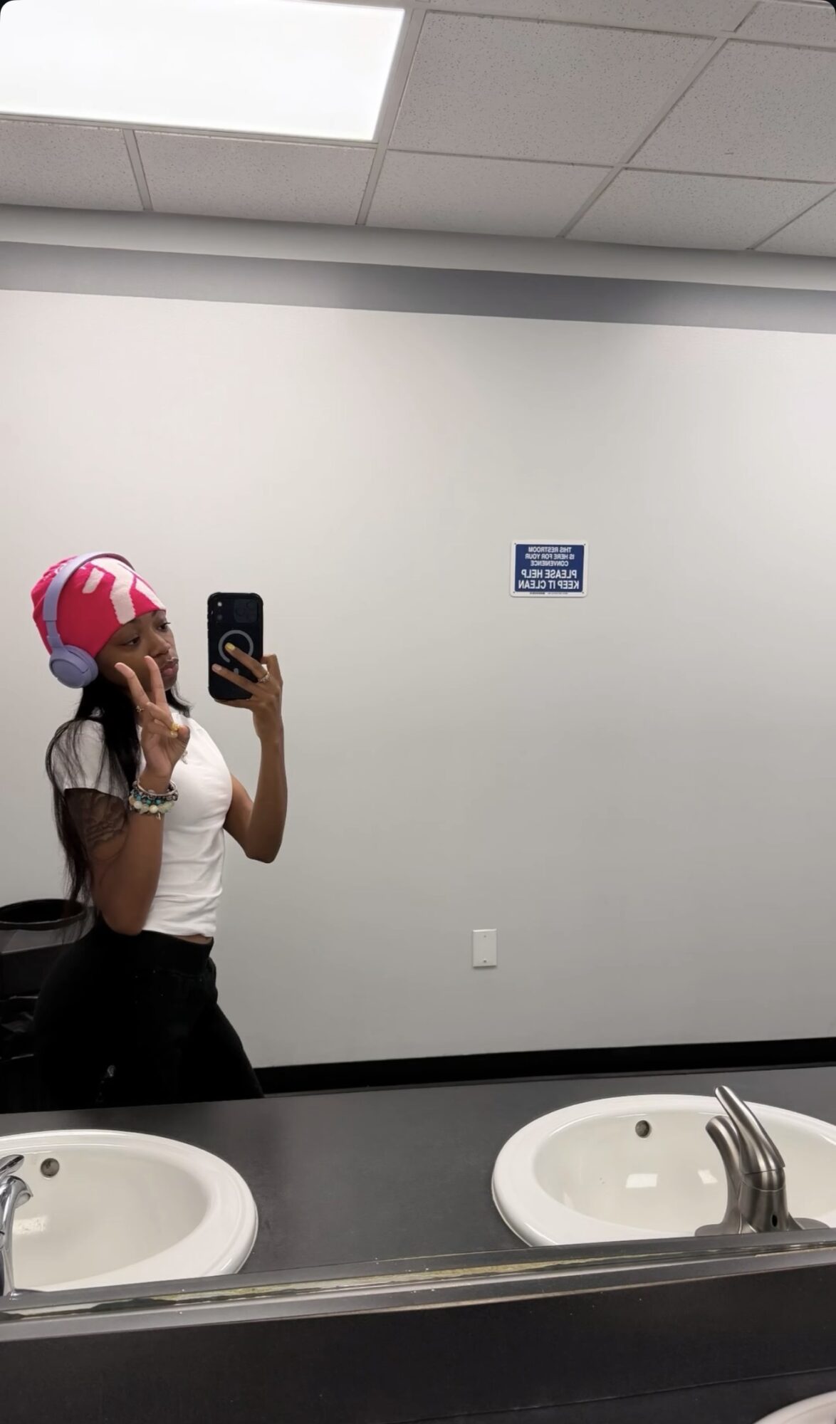 Person taking a mirror selfie in a bathroom with two sinks, wearing a pink and white striped hat and white shirt.