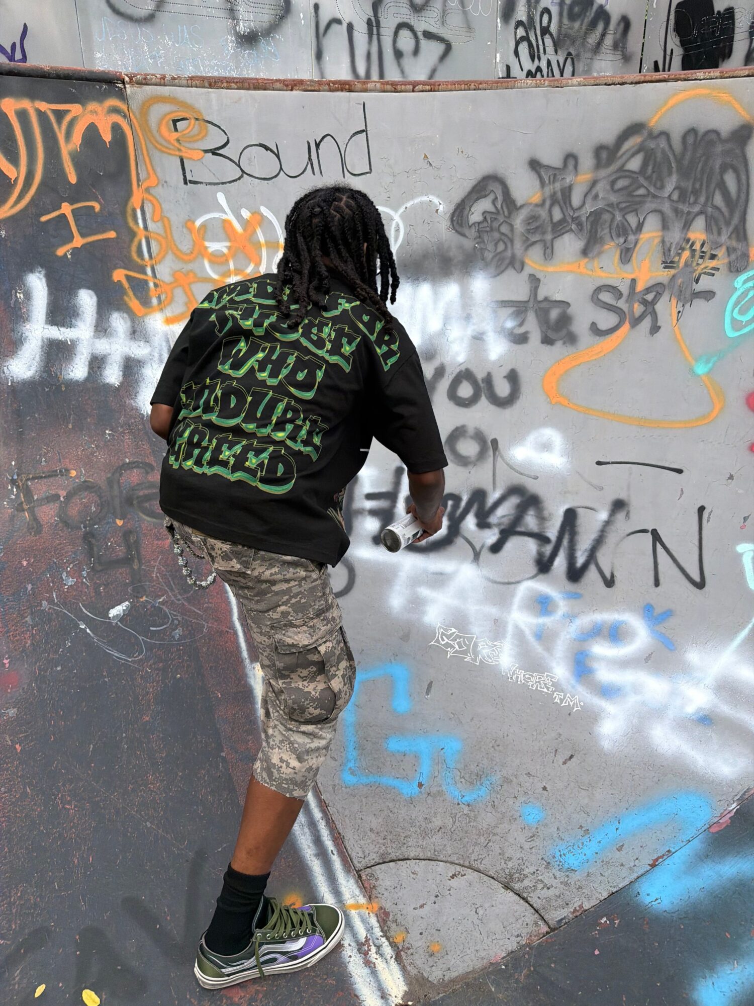 Person walking past graffiti-covered wall, wearing a black hoodie with green text, camouflage shorts, and sneakers.