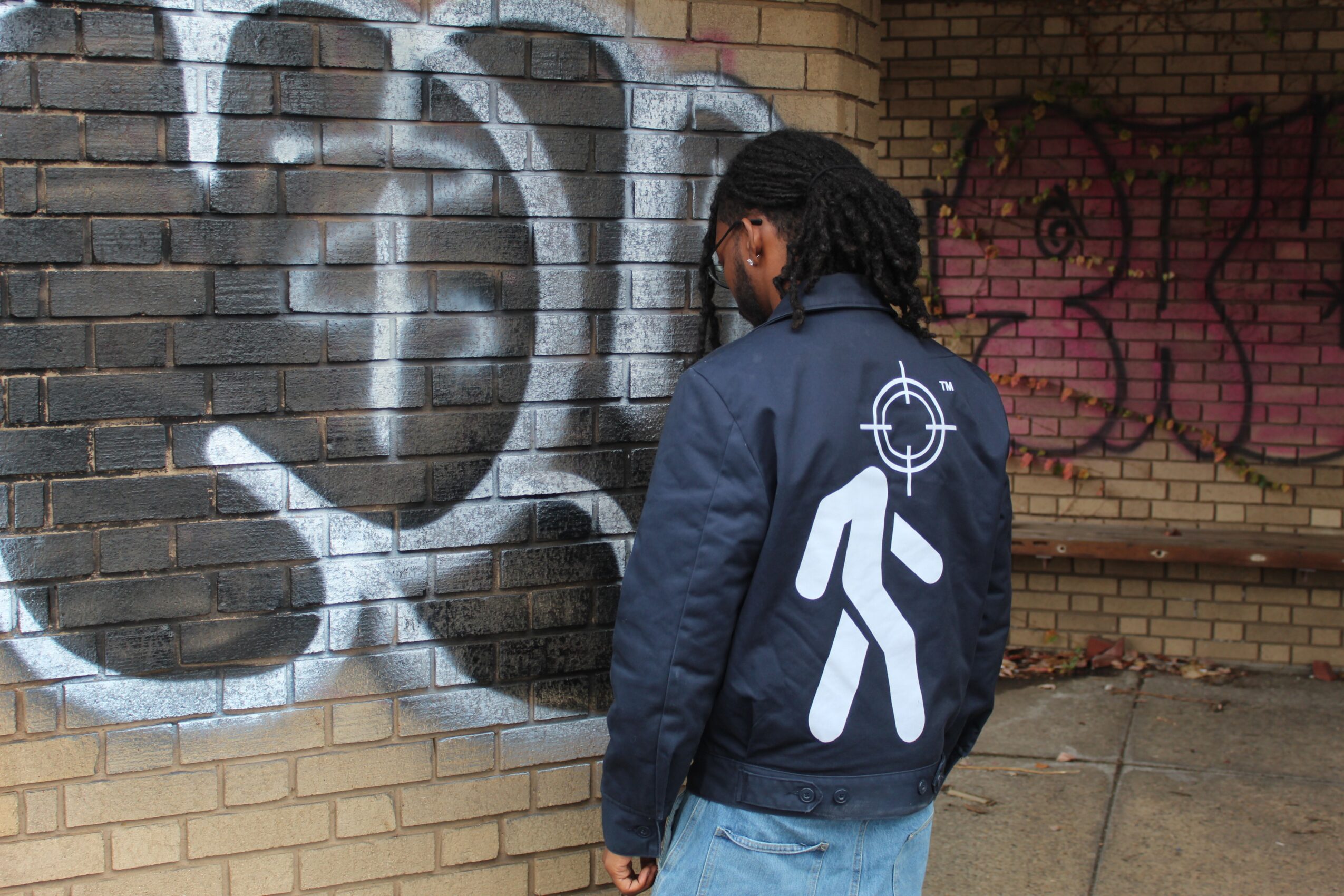 Person with dreadlocks wearing a jacket with a walking figure and crosshair symbol, standing in front of graffiti on brick wall.