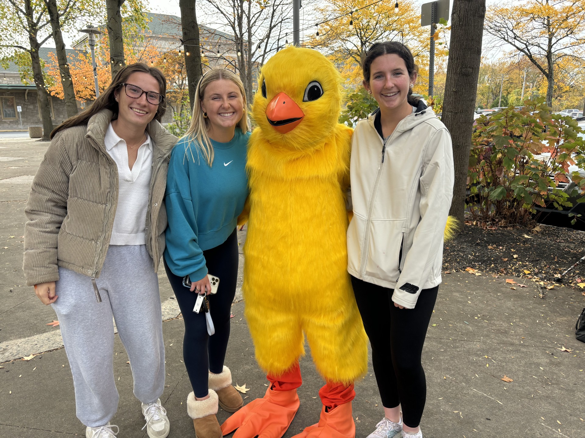 Three women standing outdoors with a person in a large yellow duck costume, trees with autumn leaves in background.