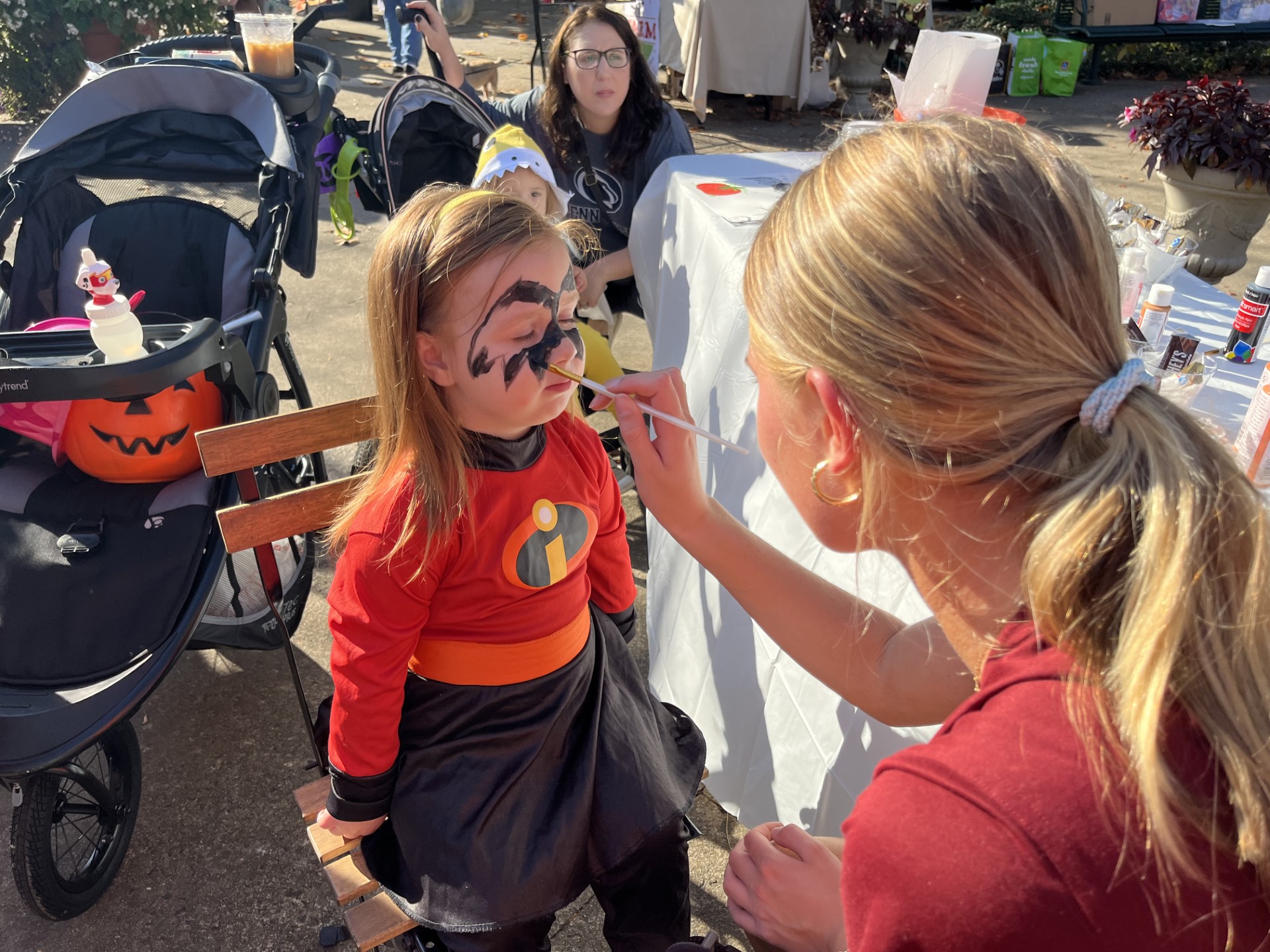 Child with face paint sitting outdoors while a woman paints her face, with a table and other people in the background.