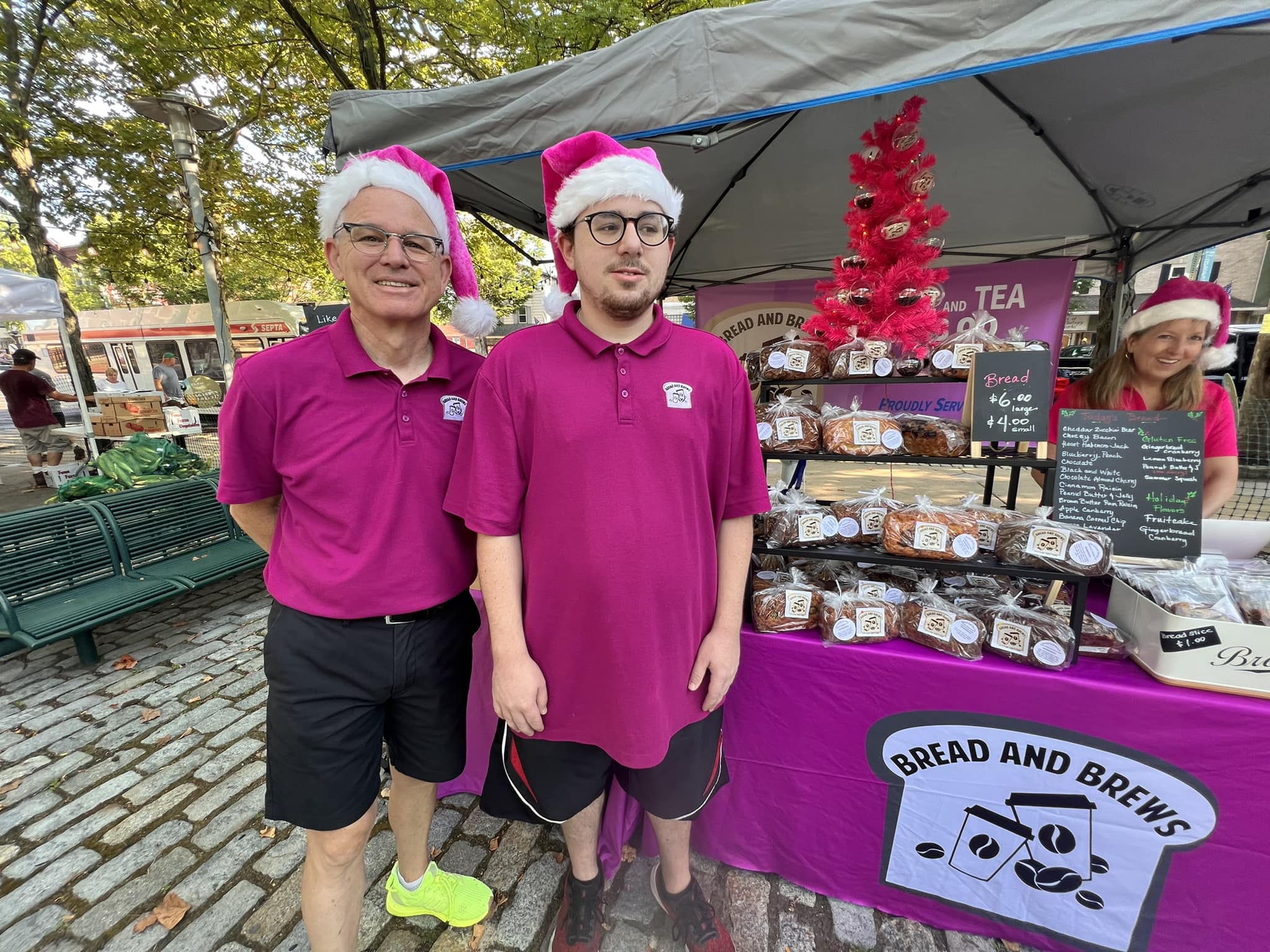 Two people wearing pink shirts and Santa hats stand in front of a pink booth with baked goods, outdoors on a cobblestone path.