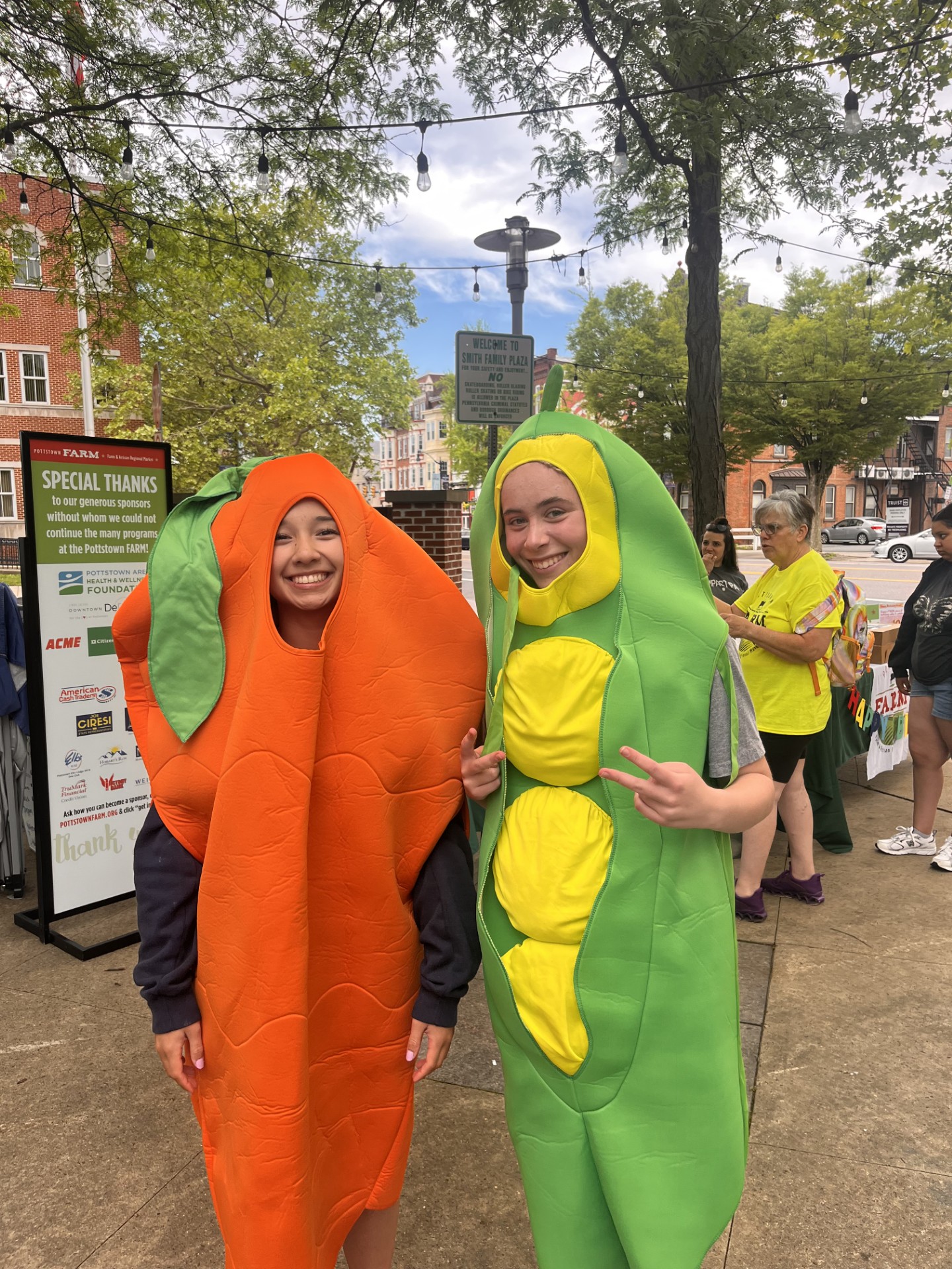 Two people in vegetable costumes, one in a carrot and the other in a cucumber, smiling outdoors on a sidewalk.