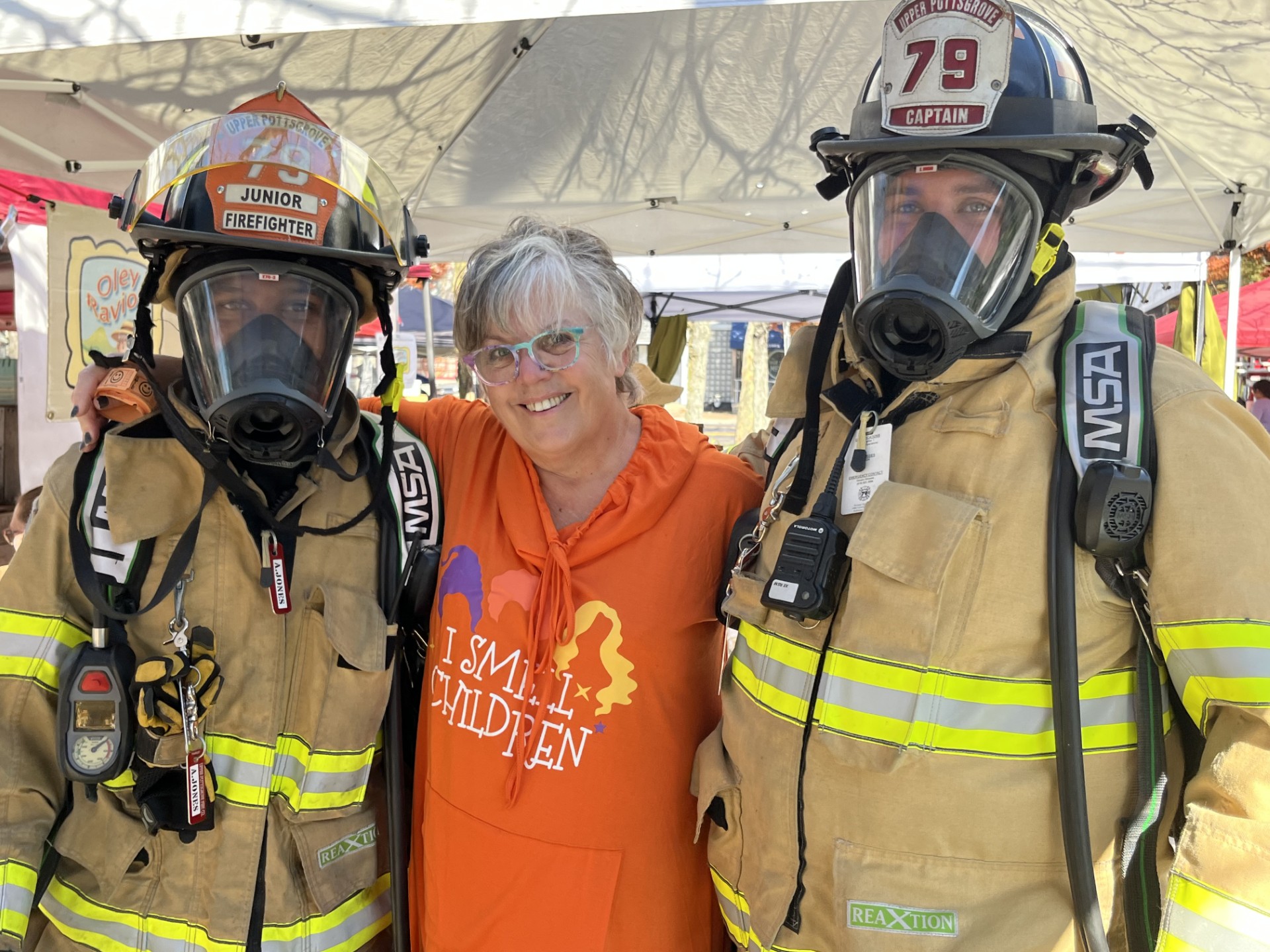 Two firefighters in protective gear stand on either side of a smiling woman in an orange hoodie, under a tent.