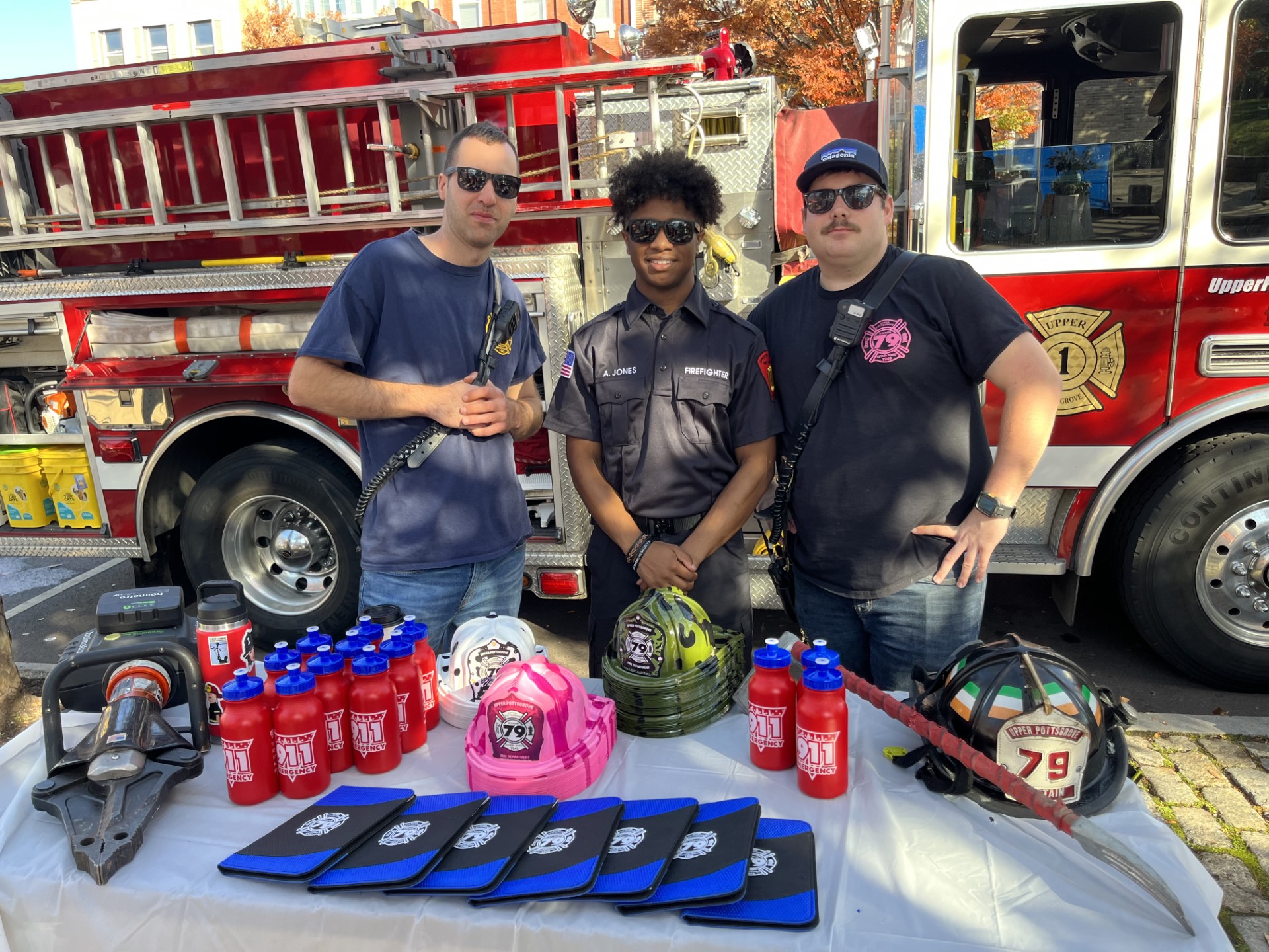 Three people stand behind a table with fire safety equipment, fire truck in background, sunny outdoor setting.