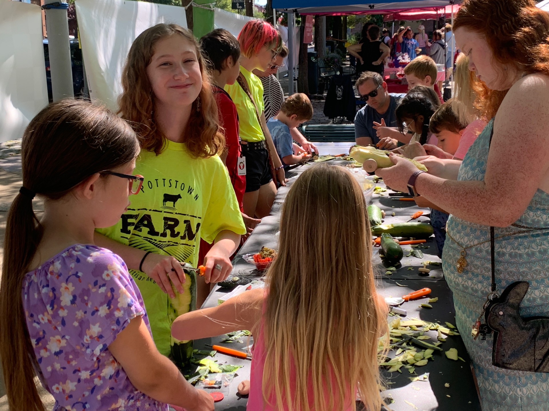 Children and adults gather around a table outdoors, engaging in a craft activity with plants and tools, sunny day.