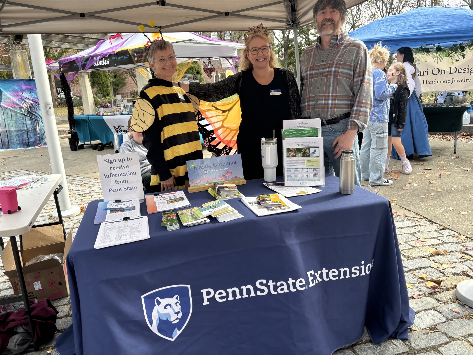 Three people stand behind a Penn State Extension table at an outdoor event, with tents and other people in the background.
