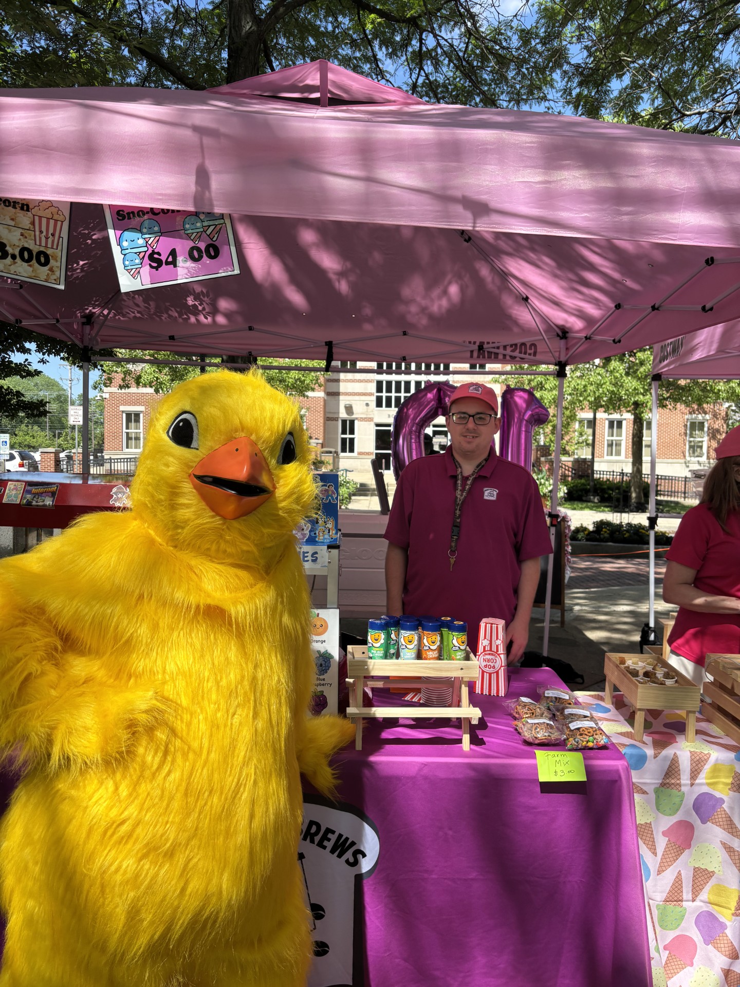 Person standing behind a booth with a pink canopy, a large yellow duck mascot in front, and various products on display.