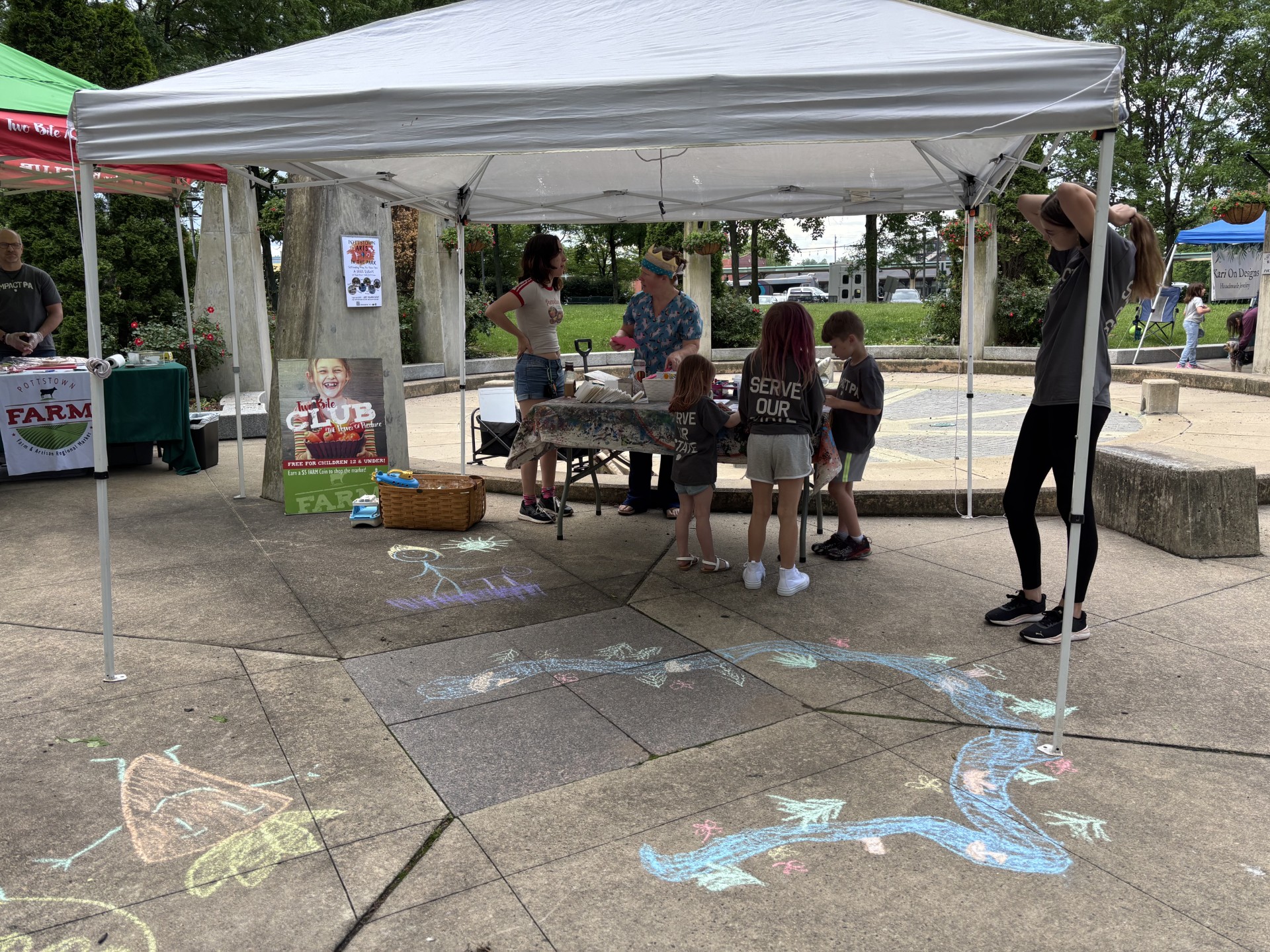 People gather under a white canopy tent at an outdoor event, with chalk drawings on the ground and trees in the background.