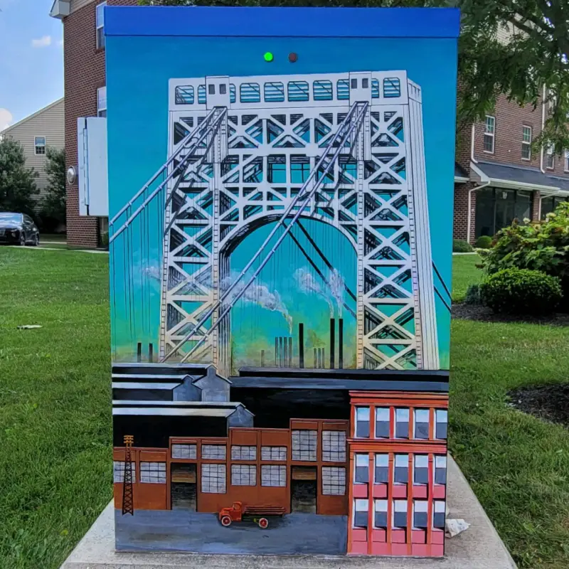 Mural of a bridge with city skyline in the background, painted on a utility box in a grassy area.