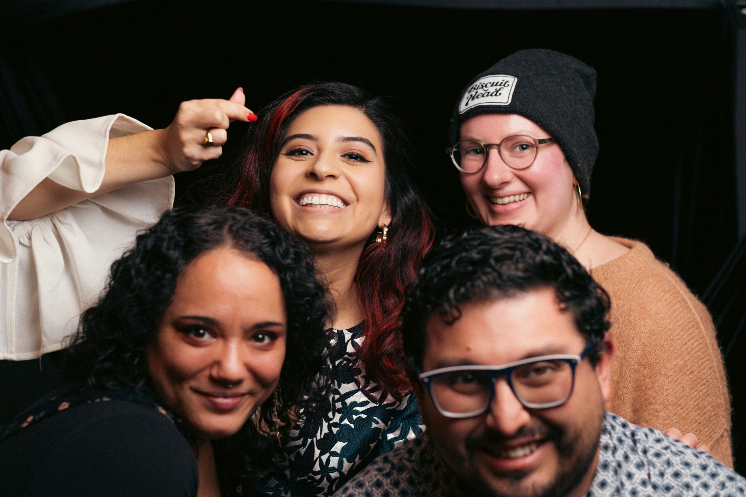 Four smiling women posing together, one with glasses and a beanie, in a dark background.