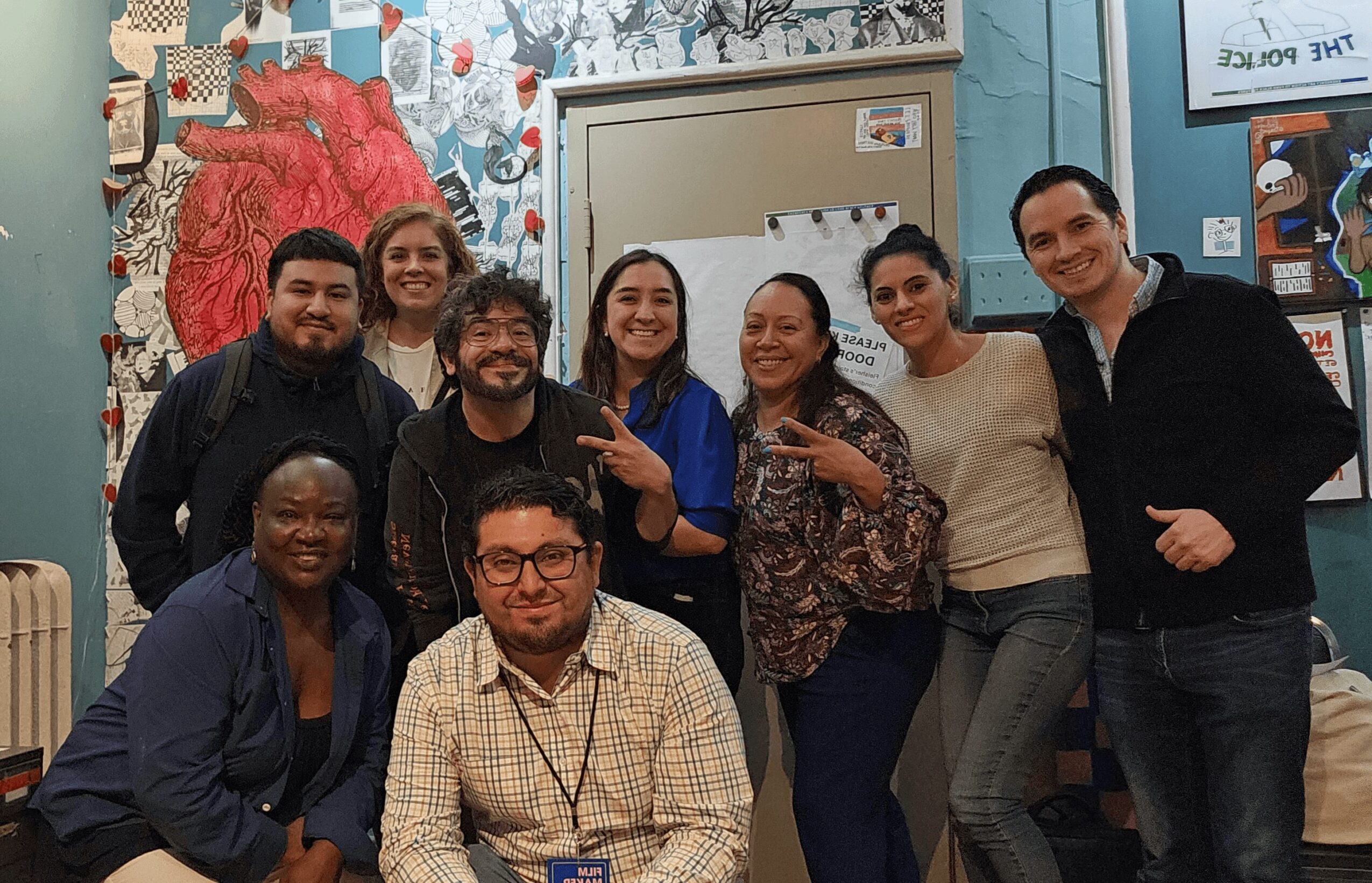 Group of nine people smiling and posing together indoors, some making peace signs, in front of colorful wall art.