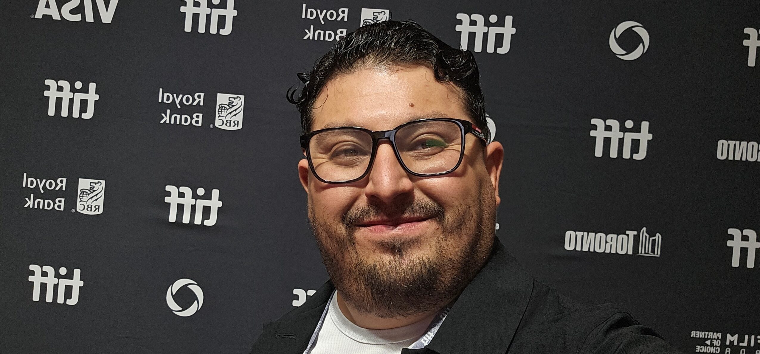Man with glasses and beard smiling in front of a black background with logos.