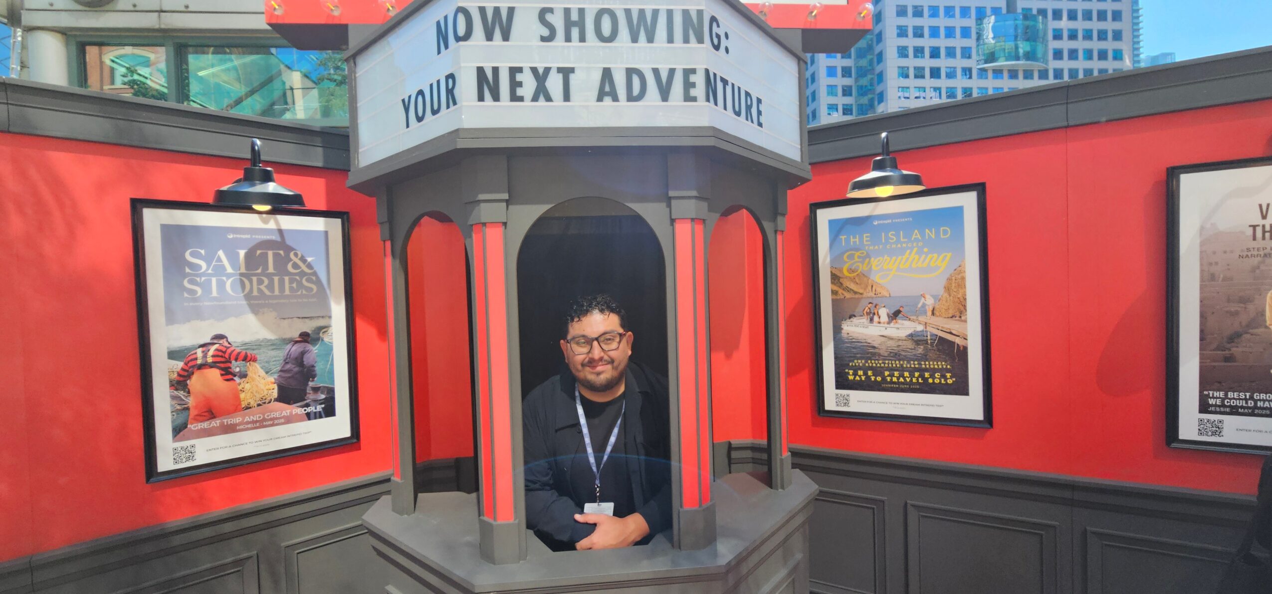 Person with glasses and dark clothing standing in front of a red theater entrance with movie posters, sign above reads 'Now Showing: Your Next Adventure'.