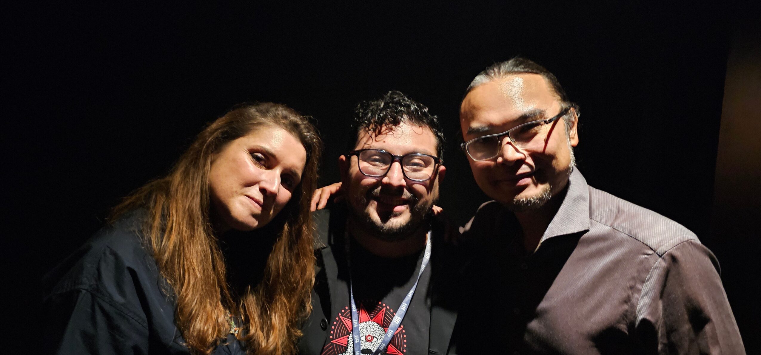 Three people smiling and posing together in a dark setting, two men and one woman, close together.