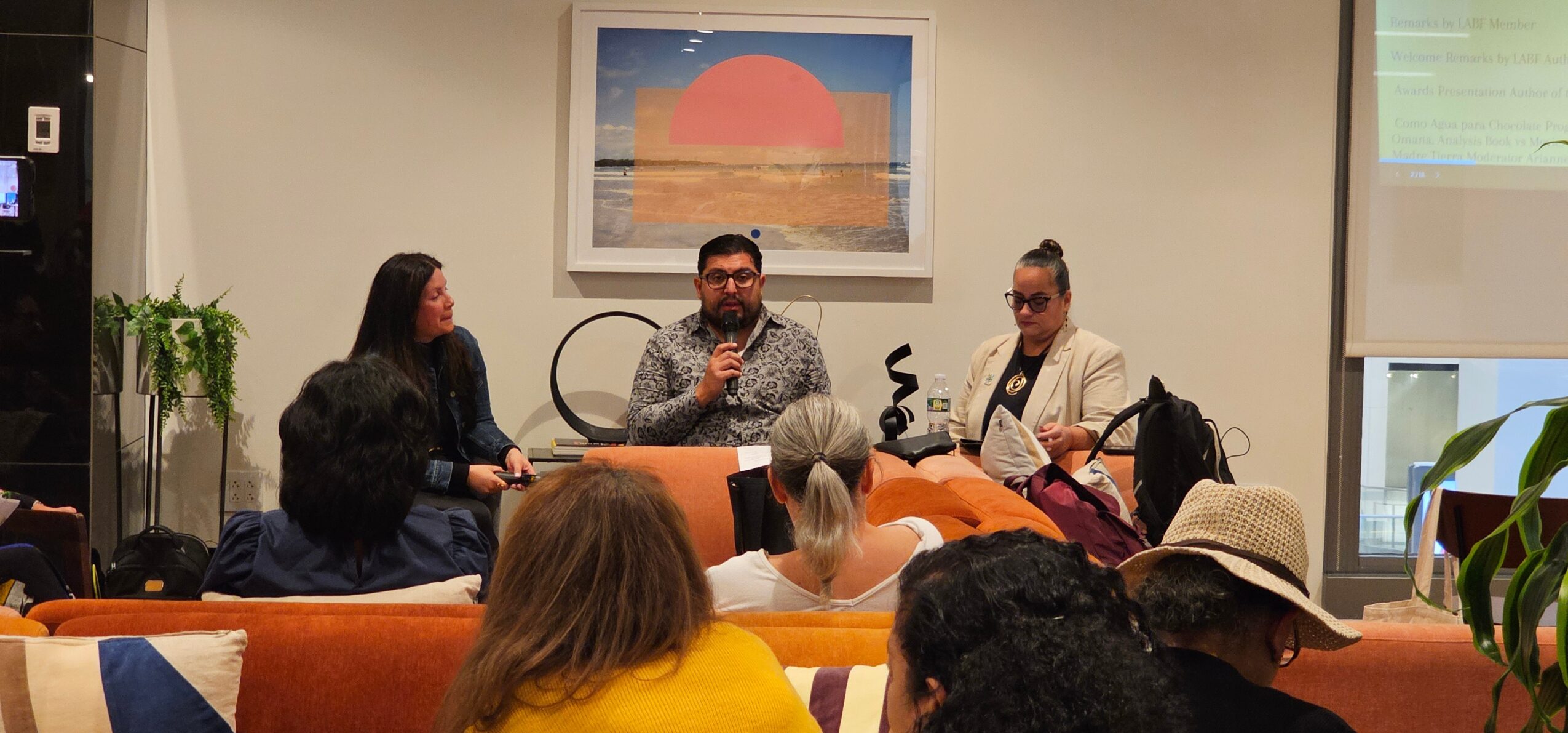 Three people seated at a panel in a room with an audience, a large colorful painting on the wall, and plants nearby.