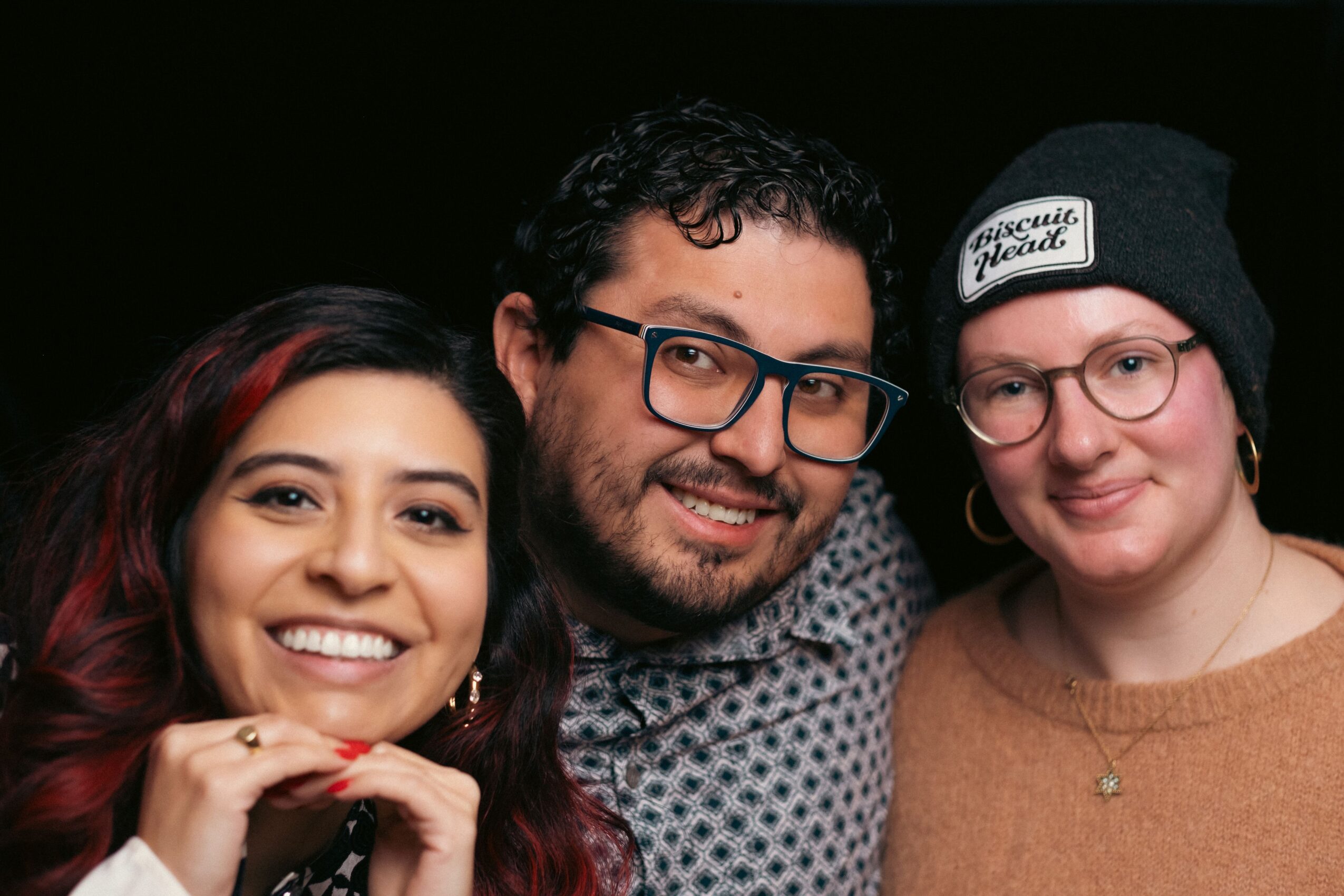 Three people smiling closely, one with glasses and a beanie, against a dark background.