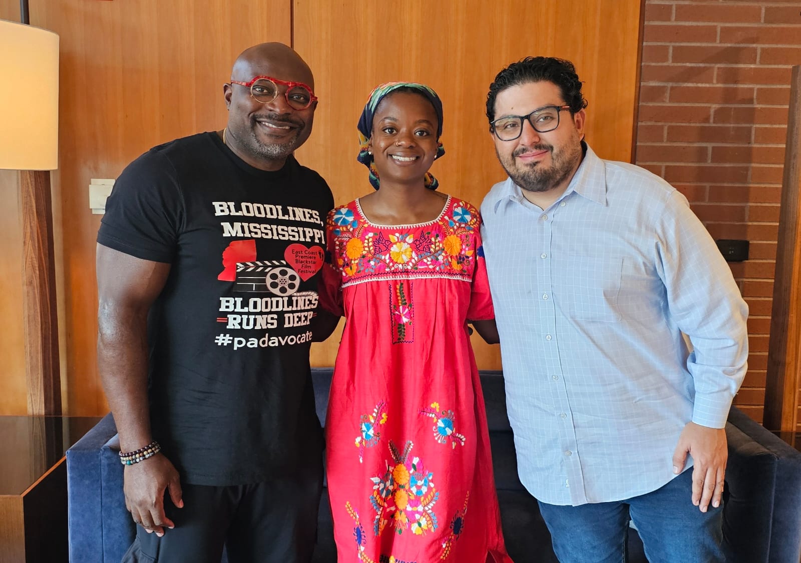 Three people standing indoors, smiling, with a wooden wall and brick wall in the background.