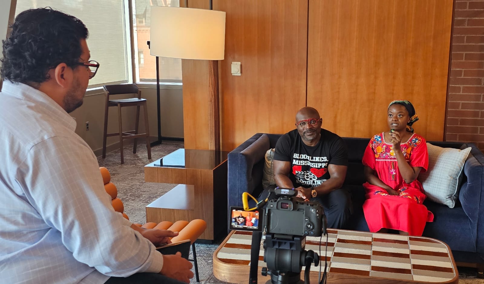 Two people sit on a sofa, being filmed by a person with a camera, in a room with wooden walls and a lamp.