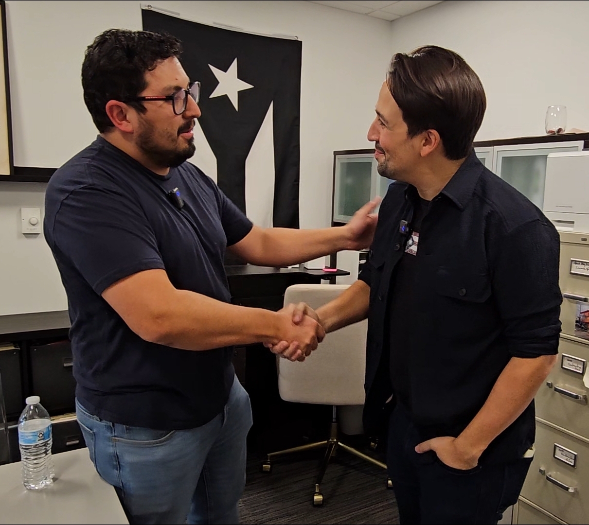 Two men shake hands and smile in an office, one with glasses and a beard, the other with dark hair and no glasses.