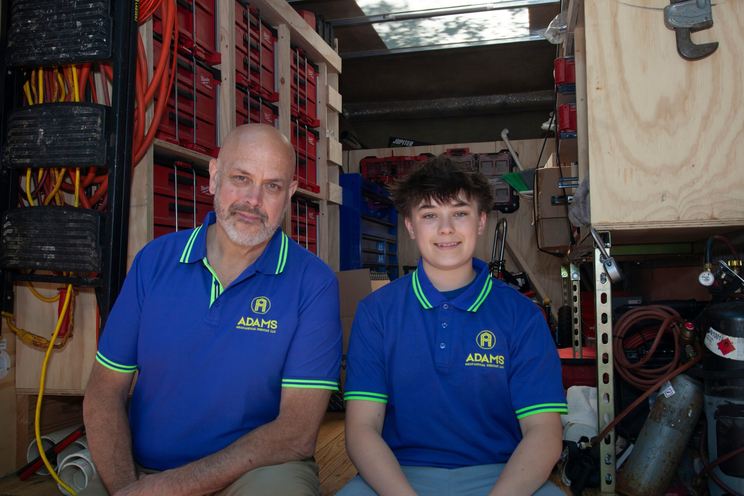 Two people in blue uniforms standing in a workshop with tools and equipment behind them.