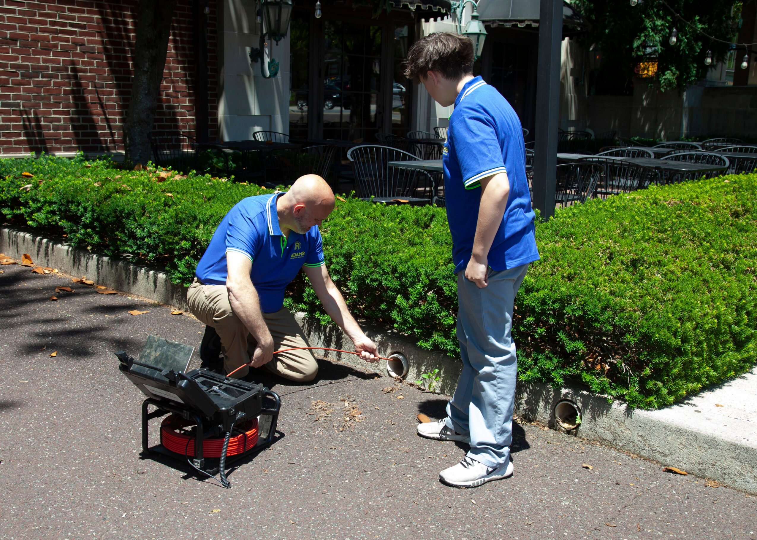 Two people working on a sidewalk near a green hedge, one kneeling and the other standing, with tools and equipment nearby.