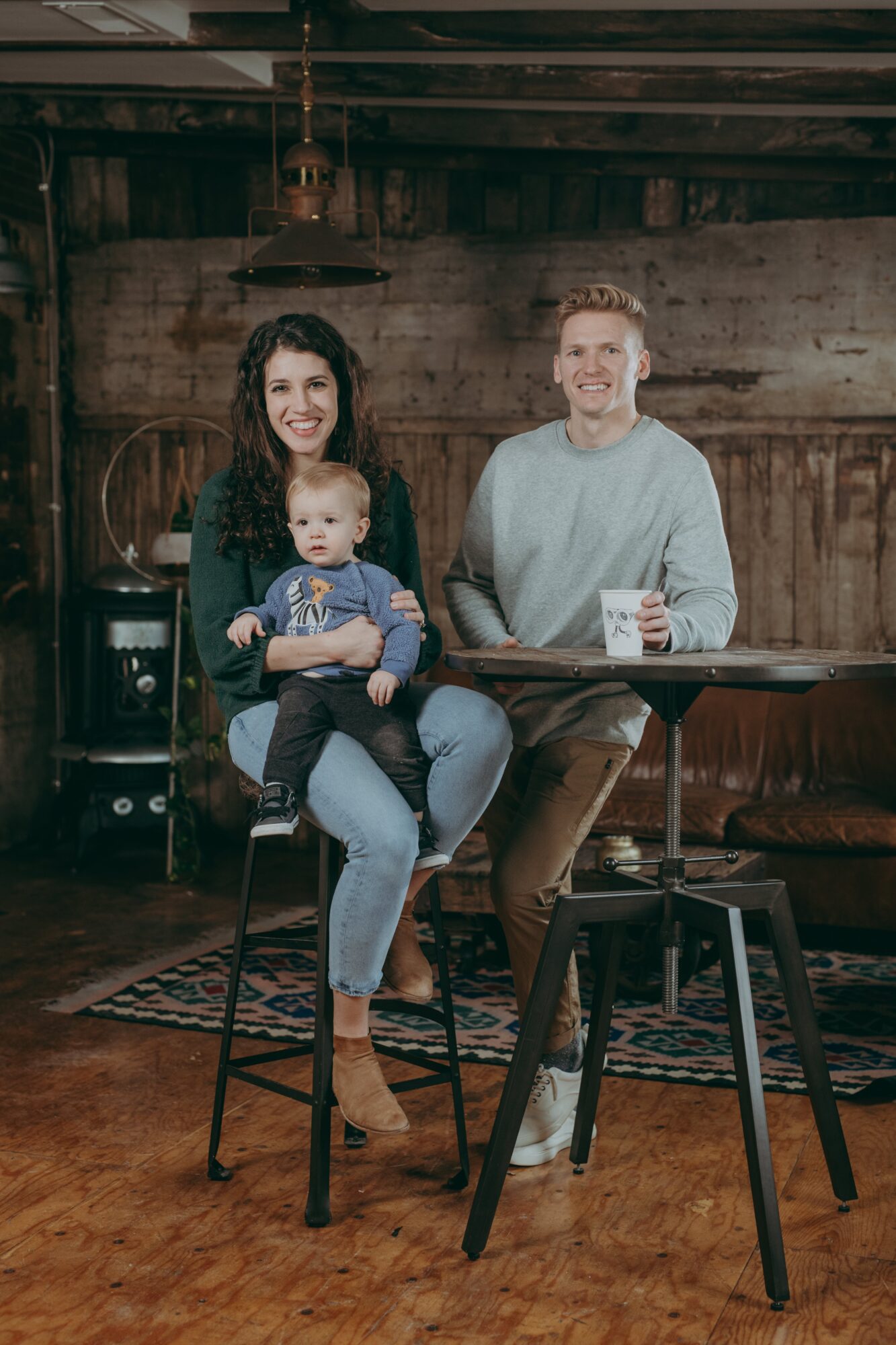 Family of three sitting in a rustic room, woman holding a baby, man sitting at a table with a cup, wooden walls.