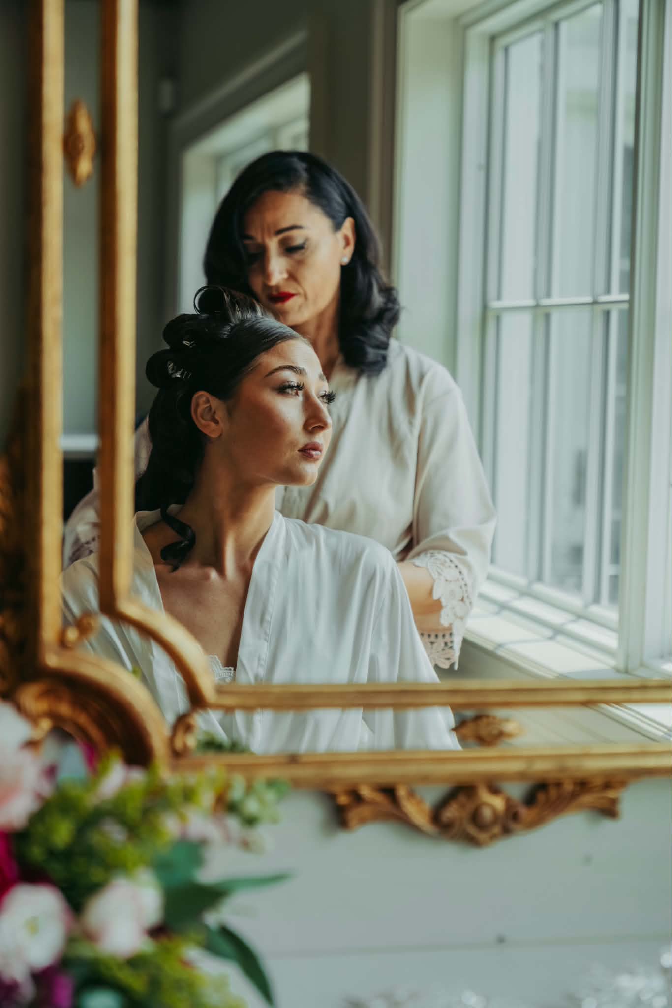 Two women with styled hair and makeup, one sitting and one standing, near a window with natural light, in a room with a mirror and flowers.