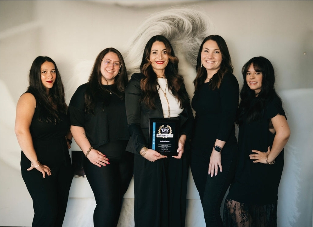 Five women standing together, one holding a plaque, smiling, in front of a plain wall with a large hair illustration.