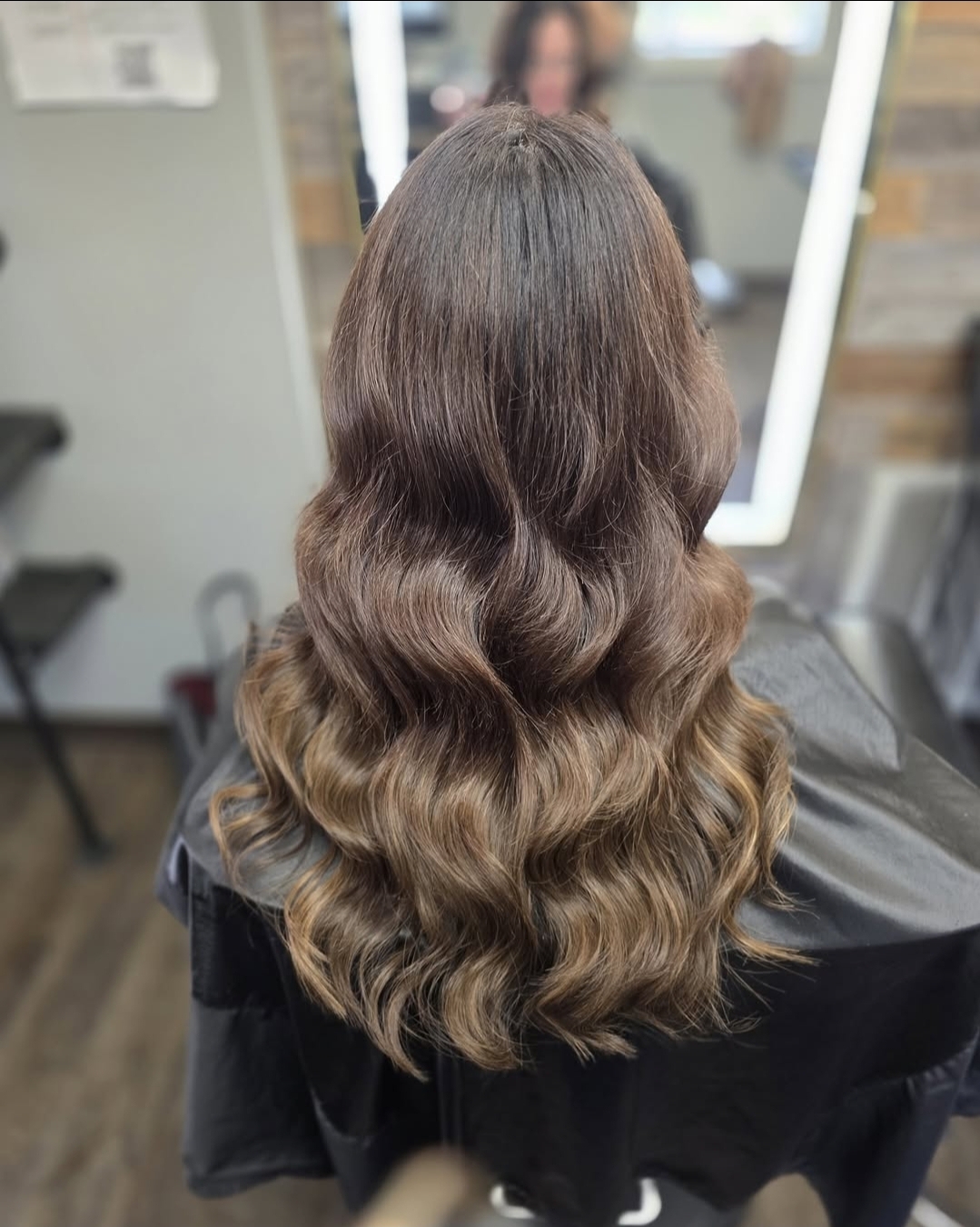 Back of a person's head with long, wavy brown hair, in a salon chair, mirror, and salon background visible