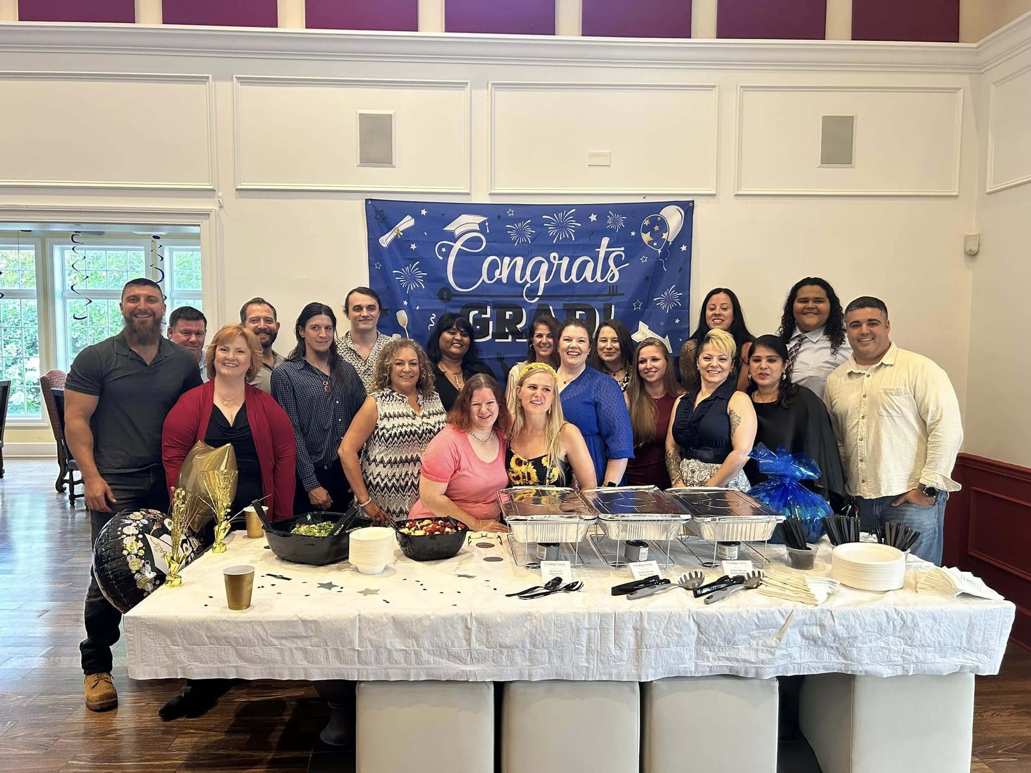 Group of people gathered behind a table with food, in front of a blue 'Congrats' banner, in a decorated room.