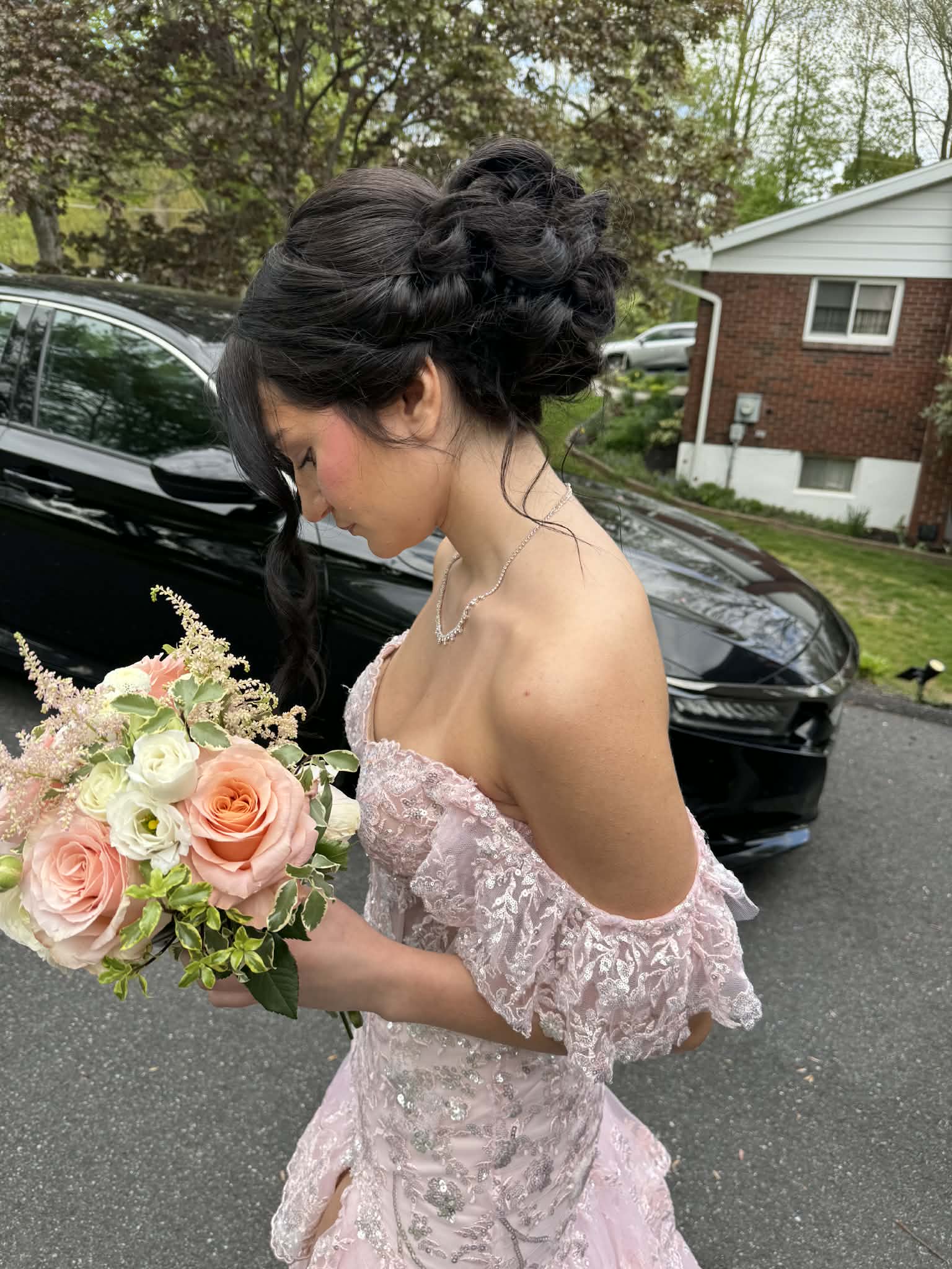 Woman with dark hair styled in an updo, holding a bouquet of pink and white roses, outdoors near a black car and house.