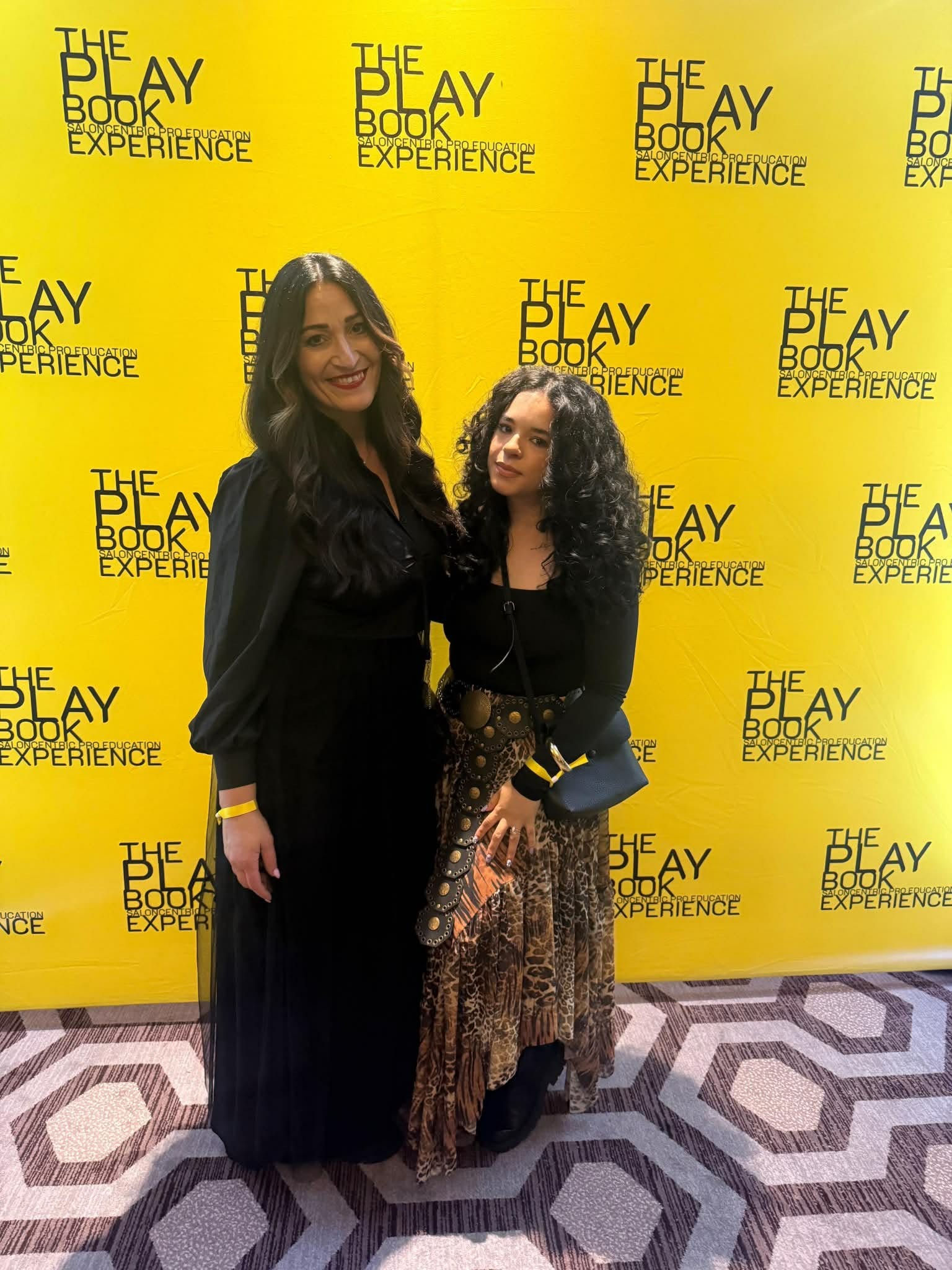 Two women standing in front of a yellow backdrop with black text, smiling, one with long dark hair, the other with curly hair.