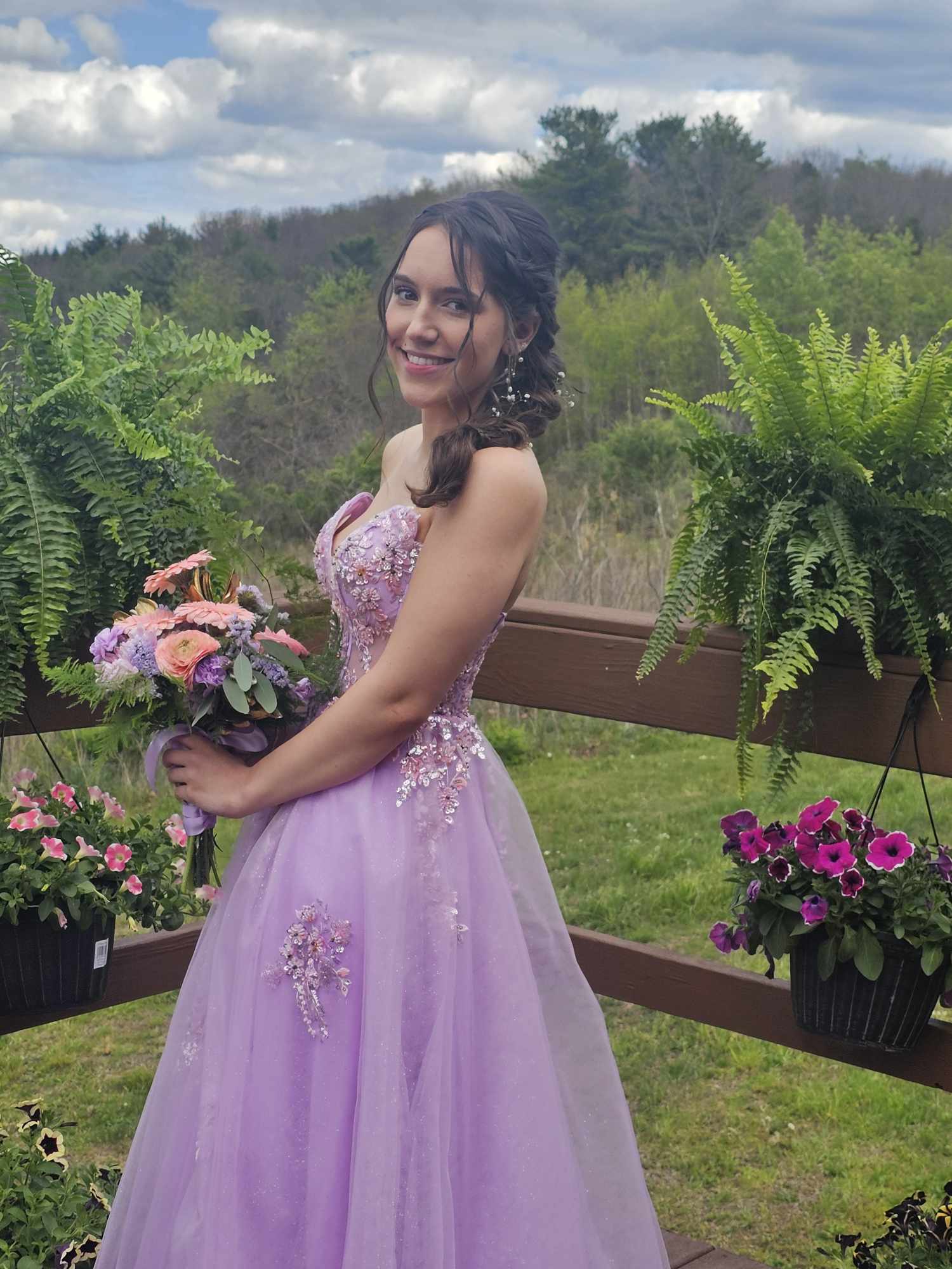Young woman in a purple dress holding a bouquet outdoors, surrounded by potted plants and greenery, smiling.
