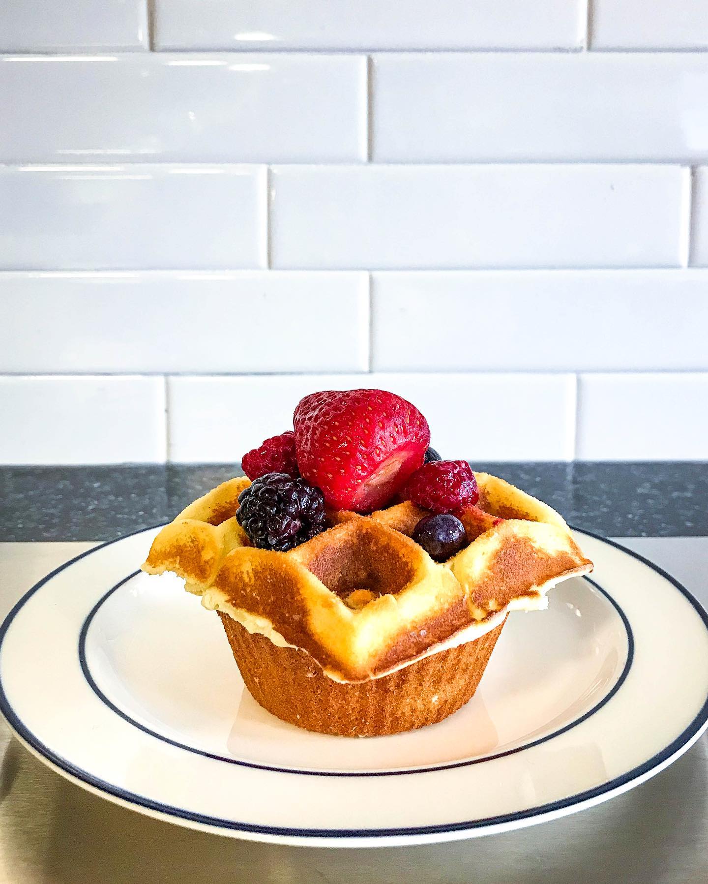 A waffle cup filled with mixed berries on a white plate with a black rim, against a white tiled background.