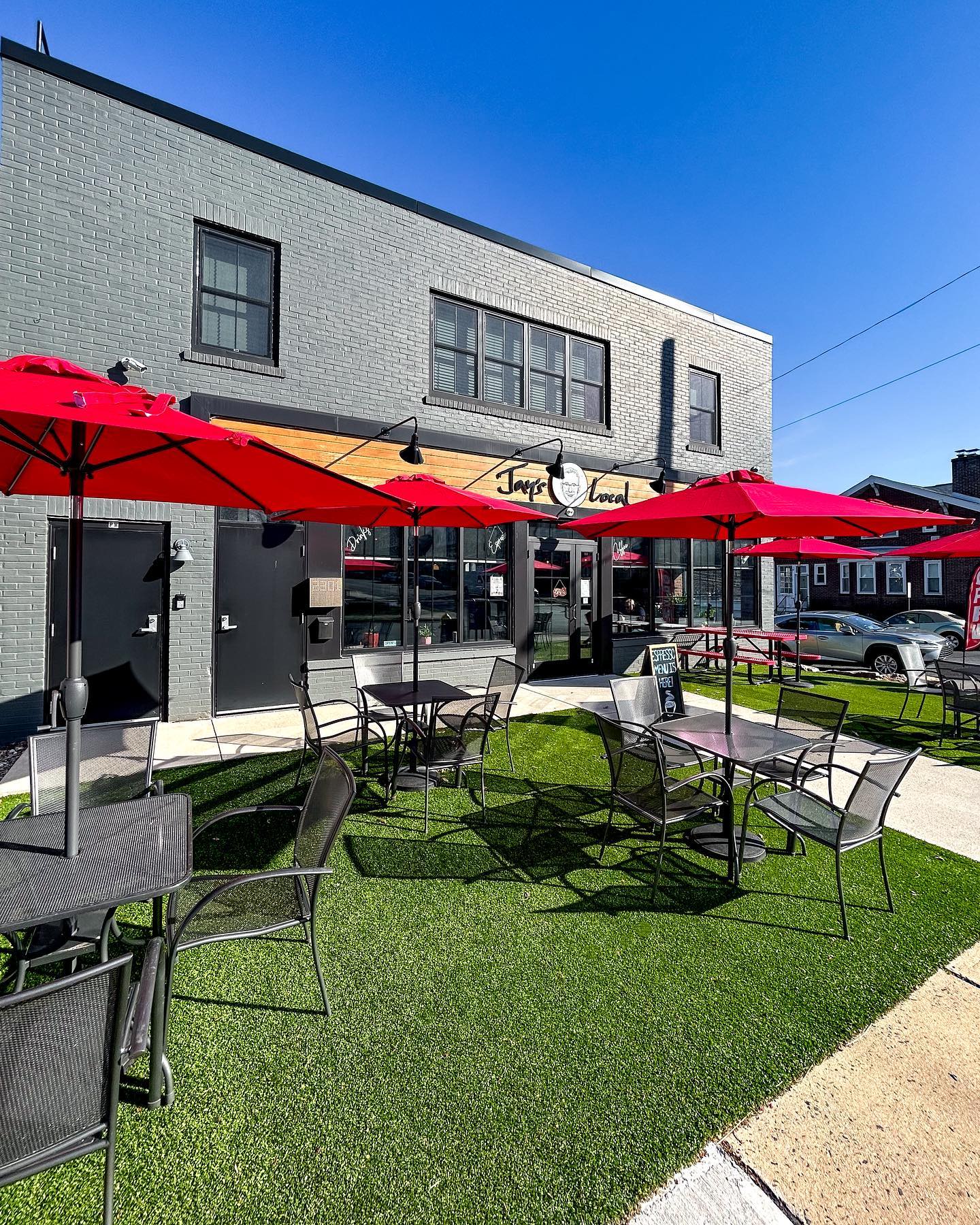 Outdoor seating area with tables, chairs, and red umbrellas in front of a two-story building under a clear blue sky.
