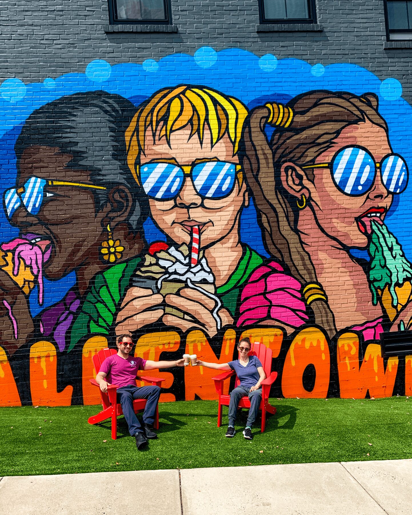 Two people sit on red chairs in front of a colorful mural of three women wearing sunglasses, with the word 'HALLOWEEN' partially visible.