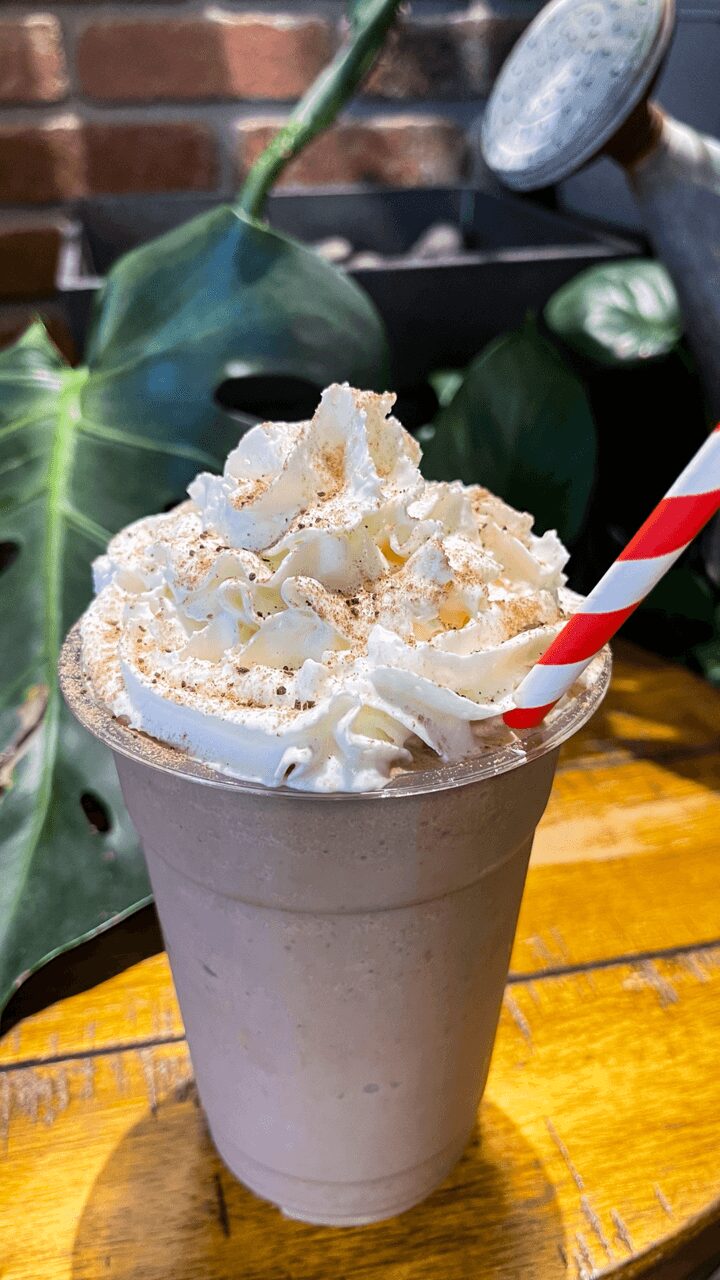 Whipped cream topping with sprinkles in a paper cup, straw, on a wooden table with green plants in background.