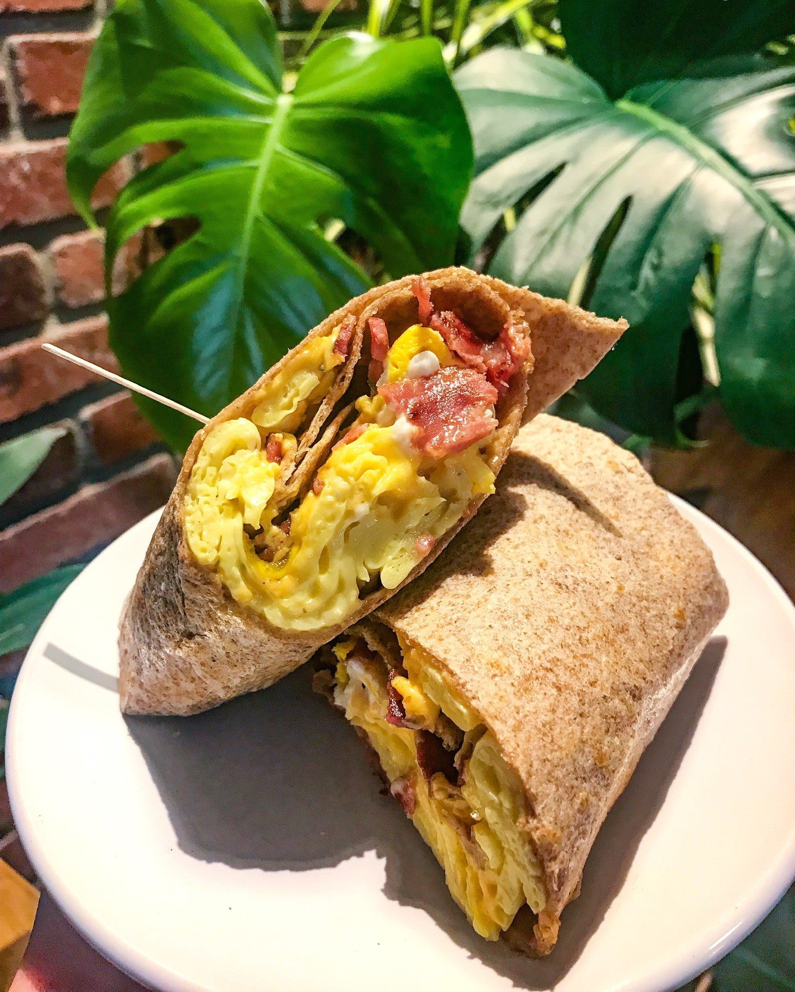 Sandwich with scrambled eggs, bacon, and vegetables on a white plate, with green plants in the background.
