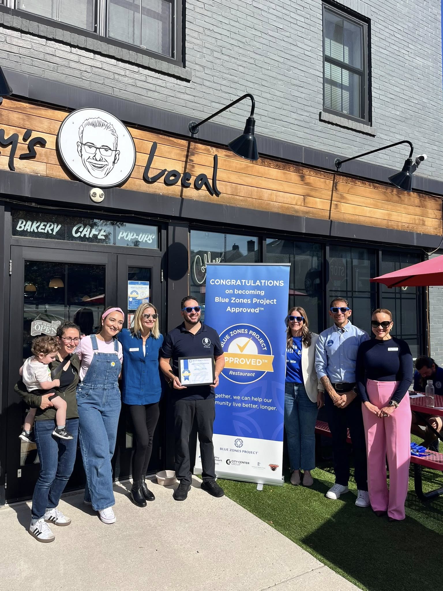 Group of people standing outside a bakery and cafe, holding a certificate, with a congratulatory banner nearby.