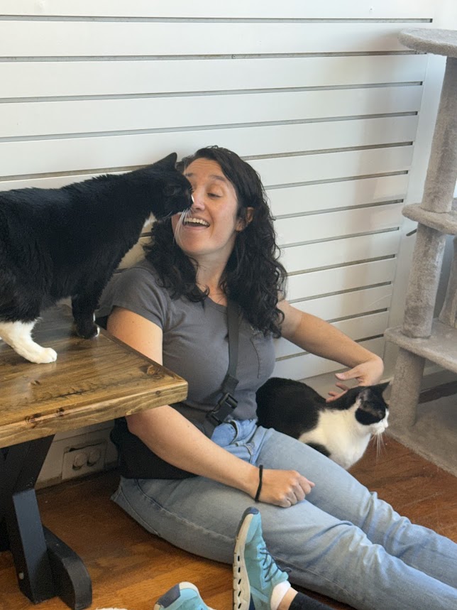 Woman sitting on the floor with two cats, one on her lap and one on a wooden table, smiling and interacting with a black and white cat. Background has white horizontal paneling.