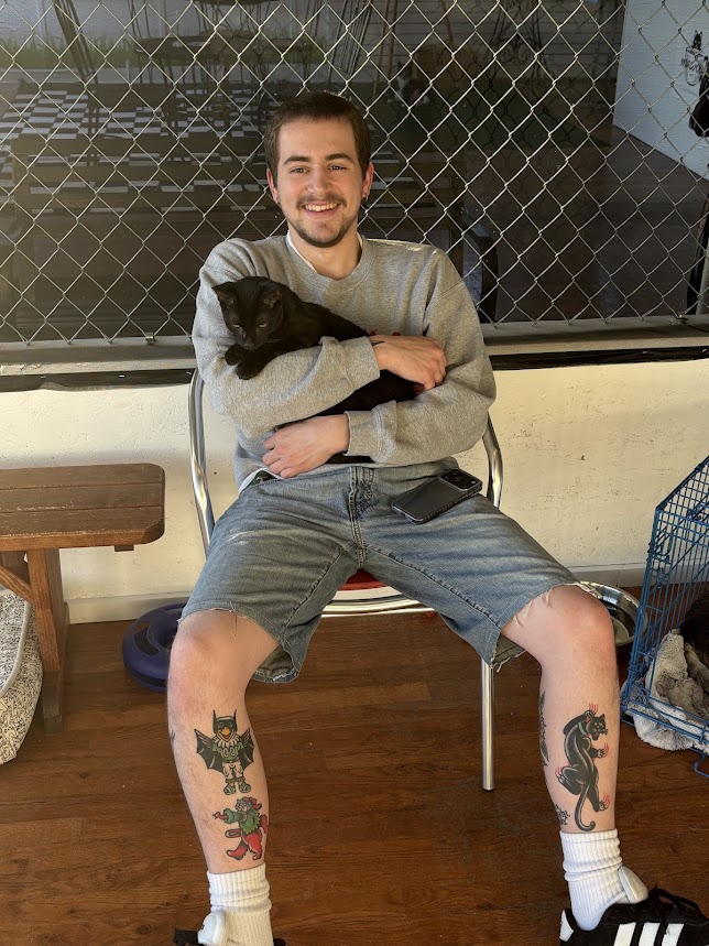 Young man with tattoos on legs, smiling, sitting on a chair holding a black cat, in a room with a chain-link fence background.