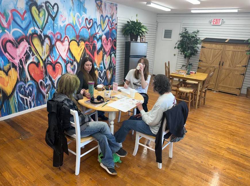Four people sit around a table in a room with a colorful heart mural, wooden floors, and potted plants.